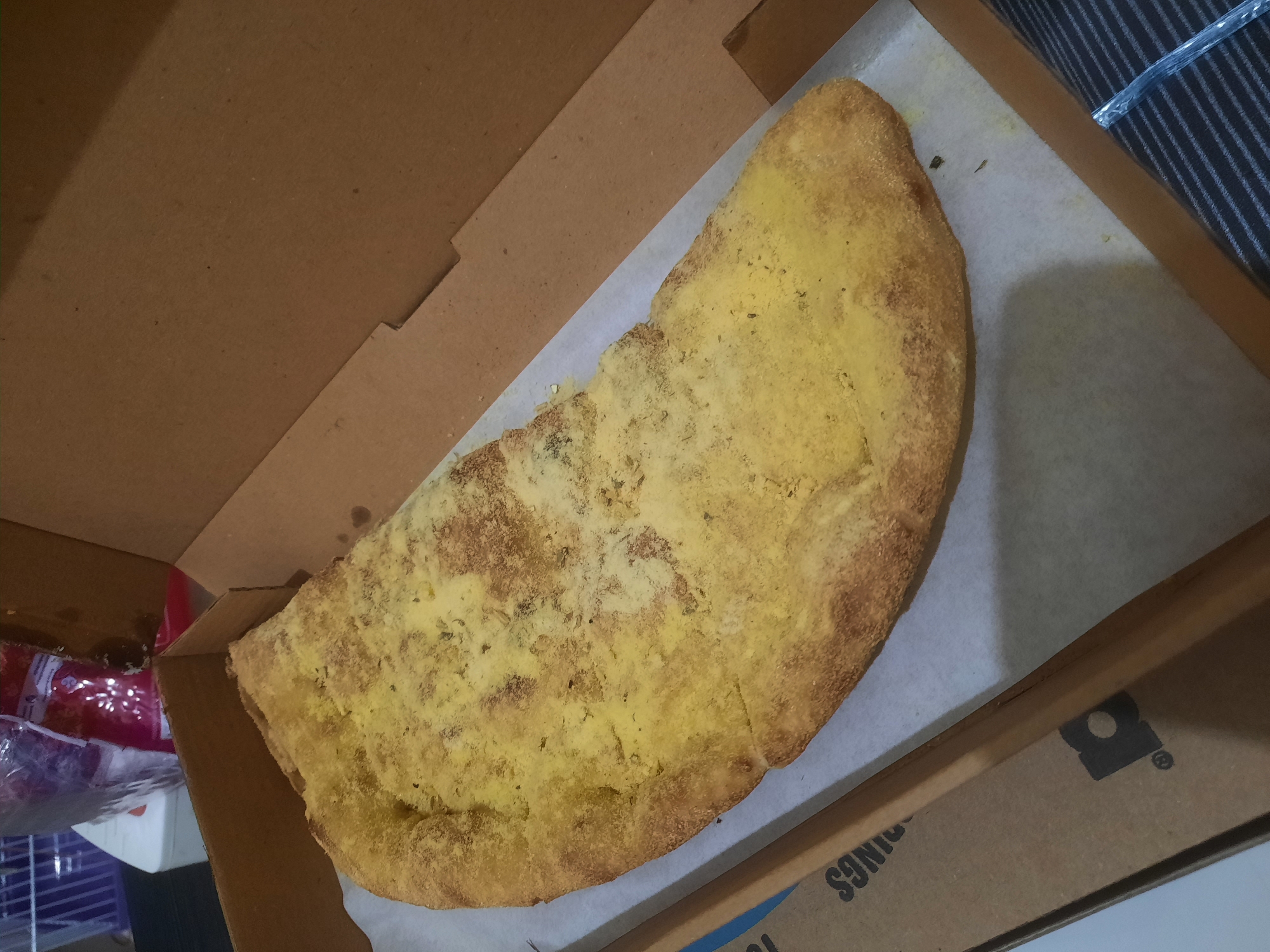 A close-up, overhead shot shows a half-moon shaped pizza with a golden-yellow topping. The pizza is presented in a brown cardboard pizza box. The crust is visible along the curved edge of the pizza, appearing golden brown and slightly puffed. The topping is somewhat unevenly distributed, with some areas appearing lighter or darker than others. Small, dark flecks are visible within the yellow topping, suggesting herbs or spices. The pizza sits on a piece of white parchment paper that lines the bottom of the pizza box. The box itself is made of corrugated cardboard and has cut-out corners. The background of the image shows a hint of a white object and some pink packaging on the left, and a dark, striped surface on the right. The setting appears to be indoors, likely a home environment, and does not contain any specific landmarks or features that would indicate a location in North Jakarta, Indonesia.