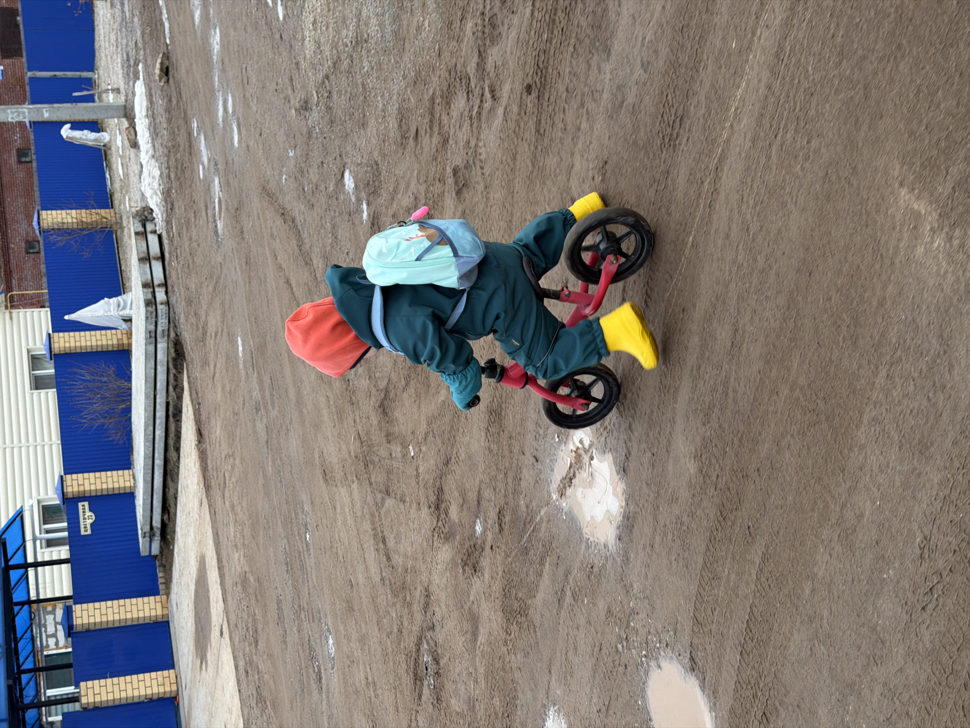 A young child in a green snowsuit, orange hat, and yellow boots is riding a red balance bike on a dirt path. The child also has a light blue backpack. The path is muddy with puddles. The location is identified as the city of Chaikovsky, Russia by a street sign visible on a blue and brick fence in the background. Patches of melting snow are present along the path and near the fence.