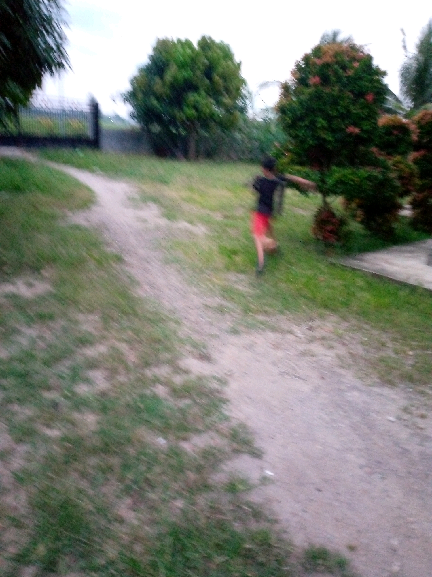 In Kutaglumpang, Indonesia, a person wearing a black t-shirt and red shorts is blurred in motion as they run along a dirt path. The path is bordered by grass and trees, including a large, leafy tree and a conical evergreen tree with red foliage. In the background, a dark metal gate is visible. The sky is overcast. The scene appears to be outdoors, possibly in a park or a residential area with ample green space.