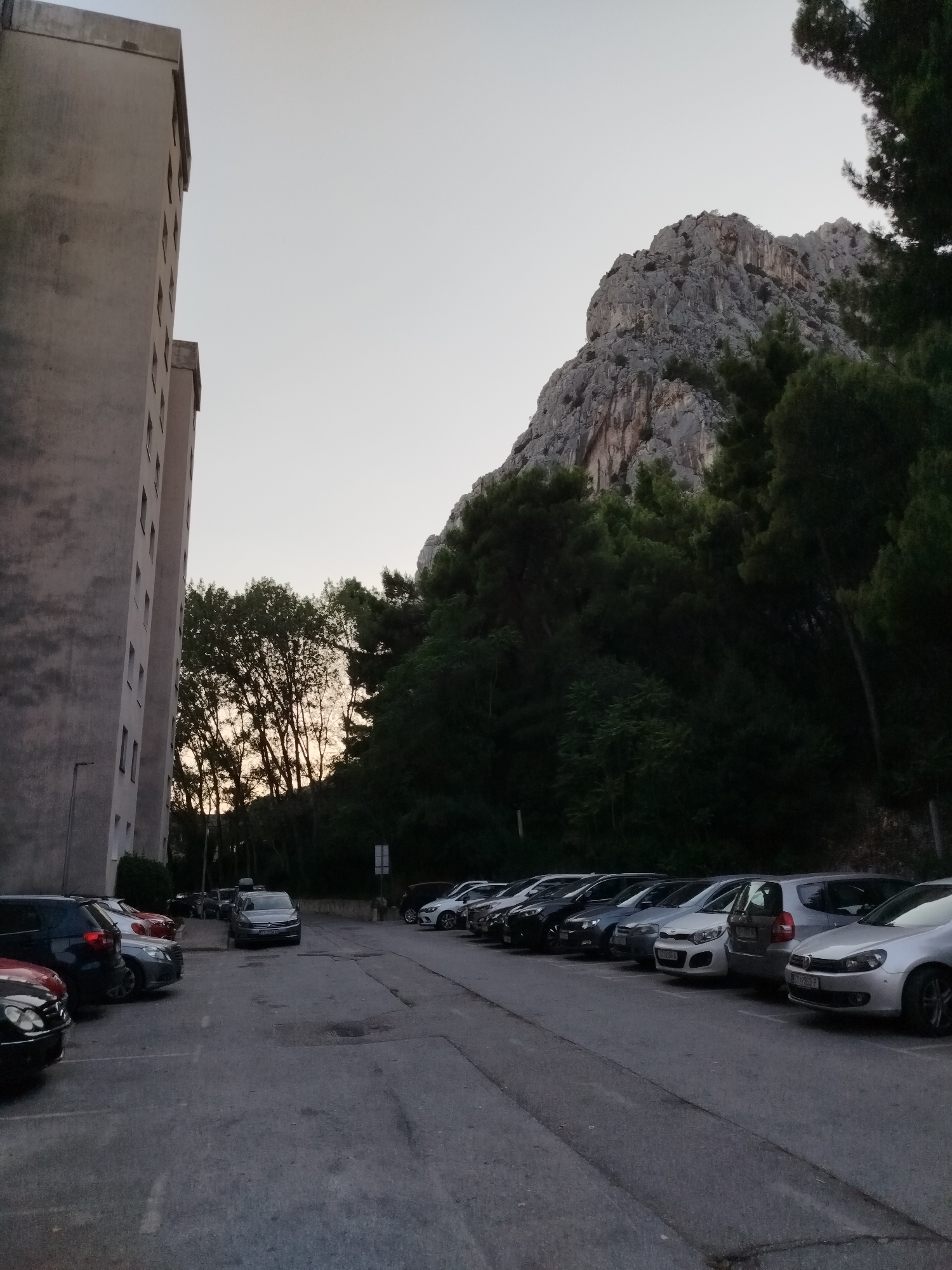The image depicts a parking lot in front of a multi-story building in Omiš, Croatia. The building, on the left side of the frame, is tall and made of light-colored concrete, with numerous windows visible. Adjacent to the building is a section of trees. In the background, a large, rocky mountain rises, partially obscured by dense green trees. A row of parked cars, including several sedans and SUVs, occupies the asphalt parking lot. The sky is a pale, overcast gray, suggesting either early morning or late evening. The overall scene is static and devoid of human activity.