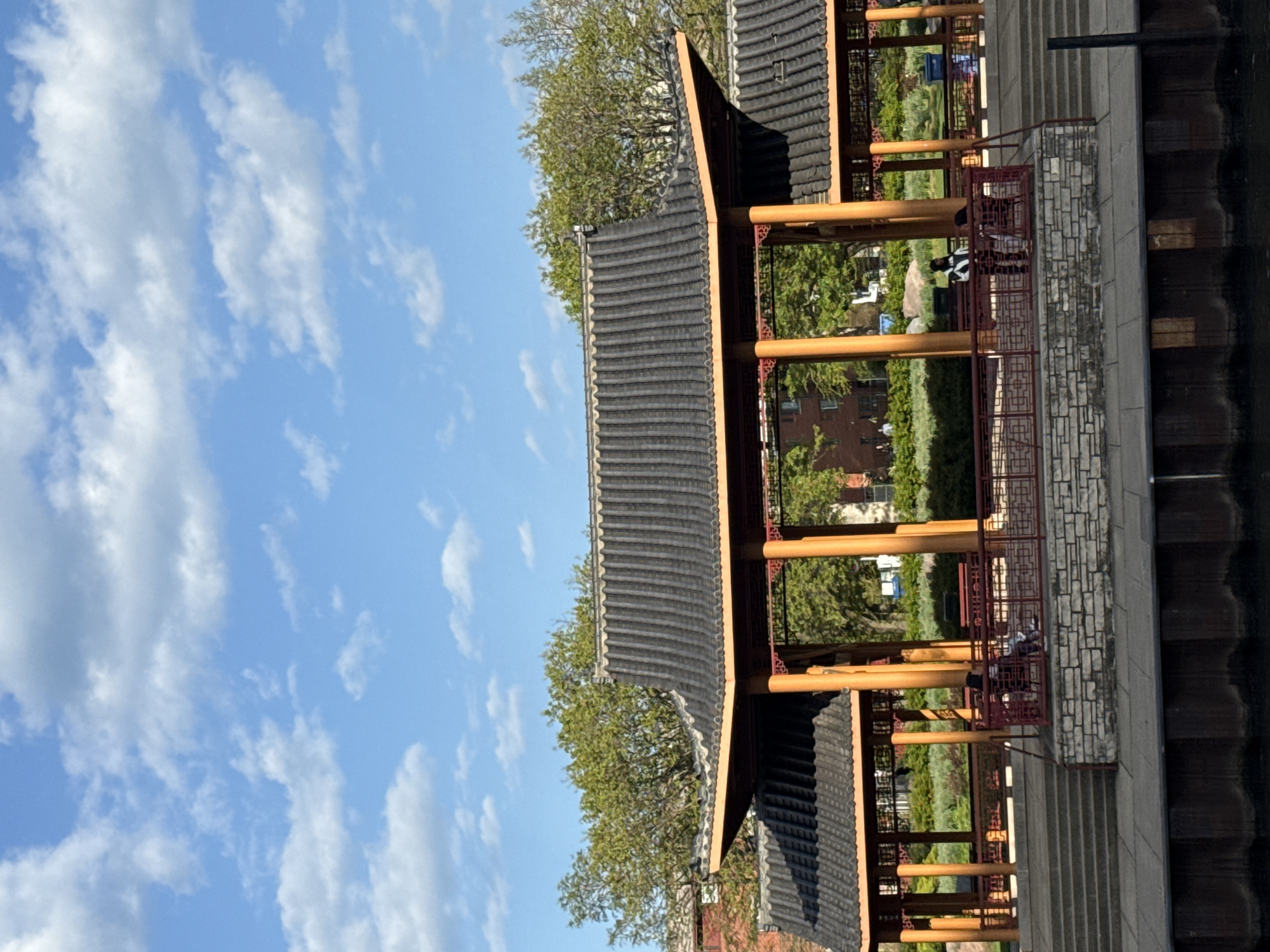 Here is a description of the image:
The image shows a structure with a traditional Chinese-style roof and wooden pillars, possibly a pavilion or pagoda. The structure is located alongside a body of water, likely the Chicago River, and is part of the Ping Tom Memorial Park in Chicago, Illinois, USA. The sky is blue with scattered clouds. There are trees and greenery visible around the structure and in the background. People can be seen on the pavilion. The image is taken from a low angle.