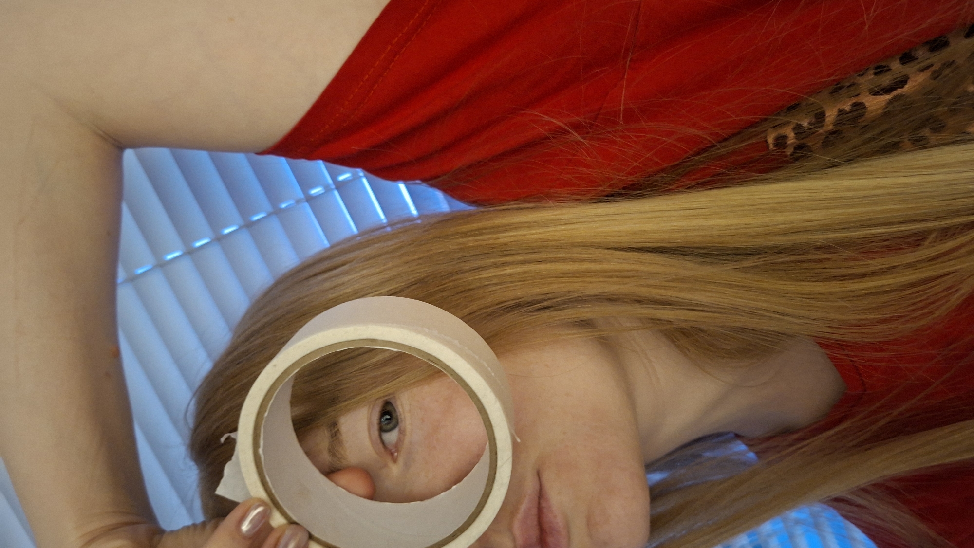 A person with long, strawberry blonde hair and fair skin is holding a roll of white tape up to their face, framing their eye. The person is wearing a red shirt. The background consists of white blinds and a small patch of fabric with a leopard print. The location of the image is not determinable from the provided image and crops. There is no indication of Chaikovsky, Russia.