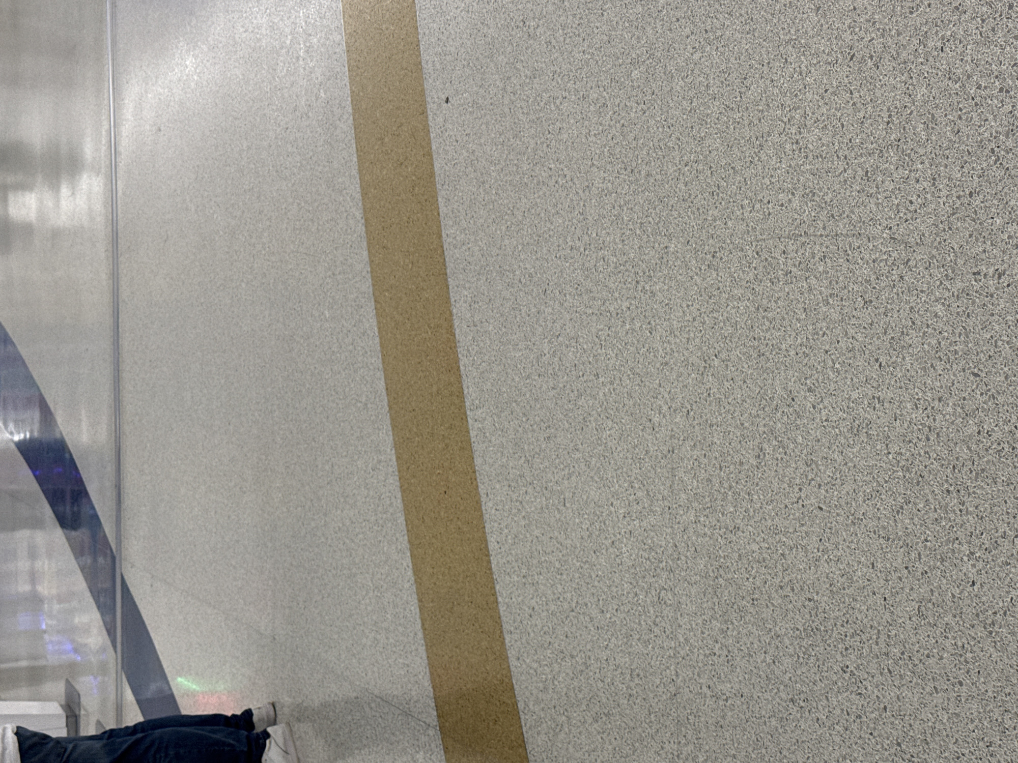 The image shows a section of flooring with a dividing gold-colored strip. To the left, there's a reflective surface with a blue stripe. In the bottom left corner, a person's lower body is visible, wearing dark-colored clothing and white shoes. Given the presence of terrazzo flooring, reflective surfaces, and the blue stripe, it's plausible that this is inside the San Antonio International Airport.