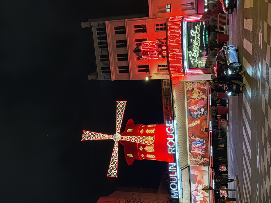 Moulin Rouge is the focus. A bright red windmill with illuminated edges takes prominence at the bottom. Above, the "Moulin Rouge" sign glows neon against the night sky. The building itself has bright posters and decor, next to the classic architecture in the background. A black car, traffic and people are visible in the street with zebra crossing.