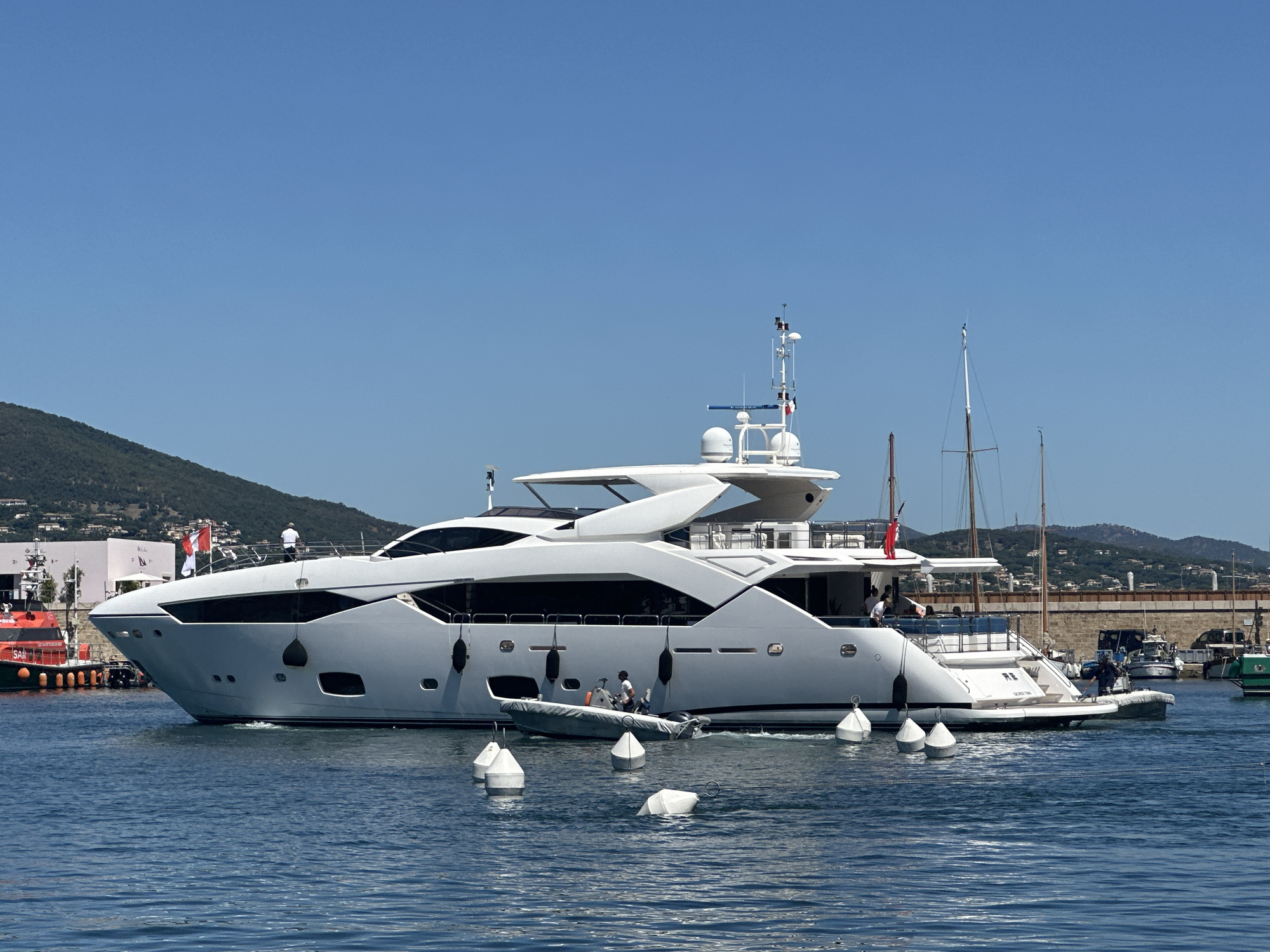 A large, white luxury yacht with multiple decks and a sleek design is moving through the water in Saint-Tropez, France. The yacht has several windows and is adorned with flags. A smaller dinghy with two people is positioned alongside the yacht. Several white buoys are floating in the blue water in the foreground. In the background, there are hills covered in vegetation, buildings, and other boats docked in a harbor. The sky is clear and blue.