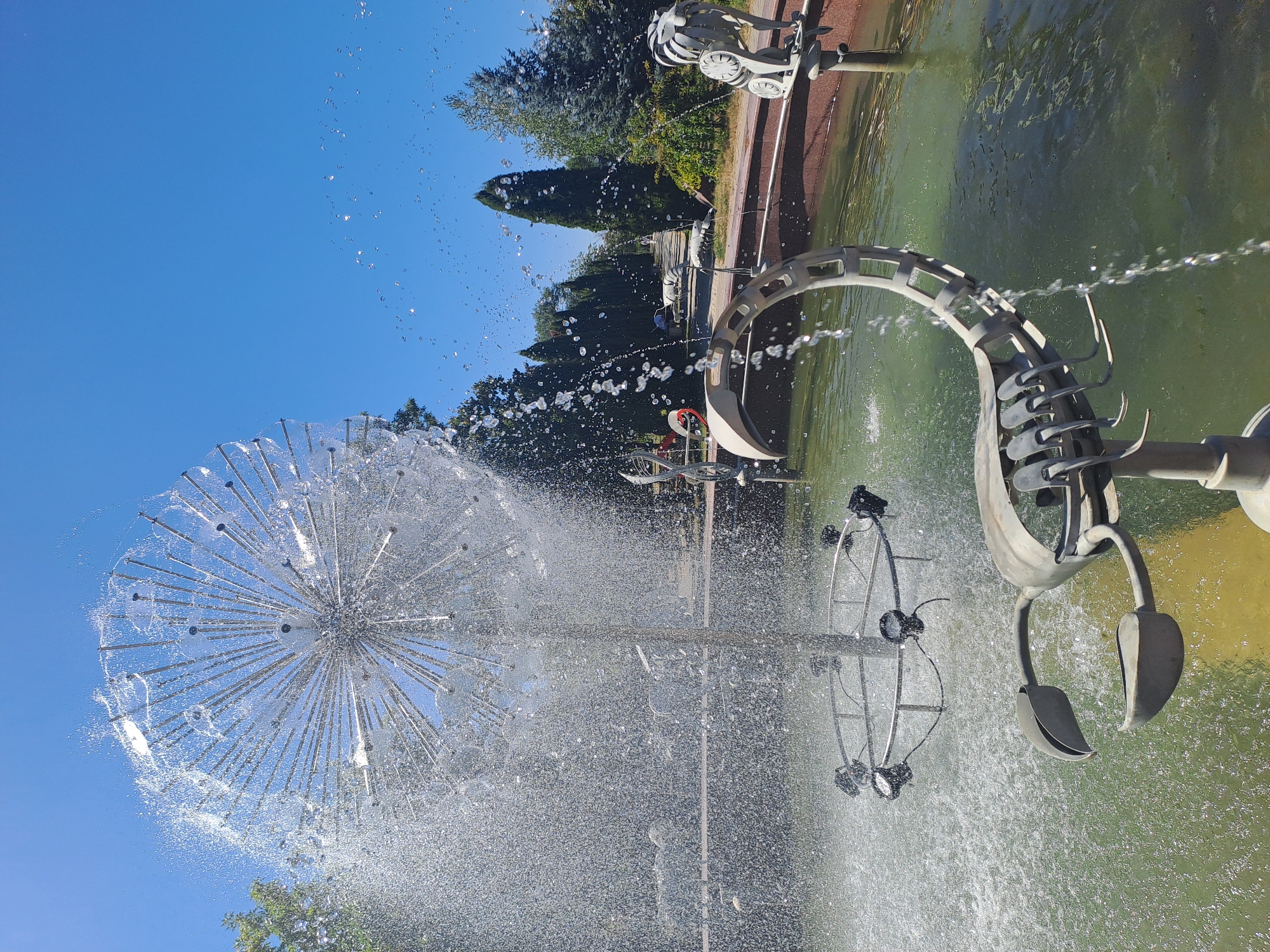 This is a daytime, eye-level, medium-shot photograph of a fountain in a park, likely in Chișinău, Moldova. The fountain features a spherical water spray, resembling a dandelion seed head, with numerous thin metal rods radiating outwards. Water is actively spraying from this structure, creating a misty effect. In the foreground, a metal sculpture of a scorpion is partially submerged in the fountain's water. The scorpion sculpture has a segmented tail that curves upwards, and its claws are open. The fountain basin is filled with greenish water. In the background, there is a paved walkway lined with cypress trees and other foliage. The sky is a clear, bright blue, indicating a sunny day. Further back, there appears to be a road with a few vehicles, and more trees. There are no people visibly present in the immediate vicinity of the fountain.