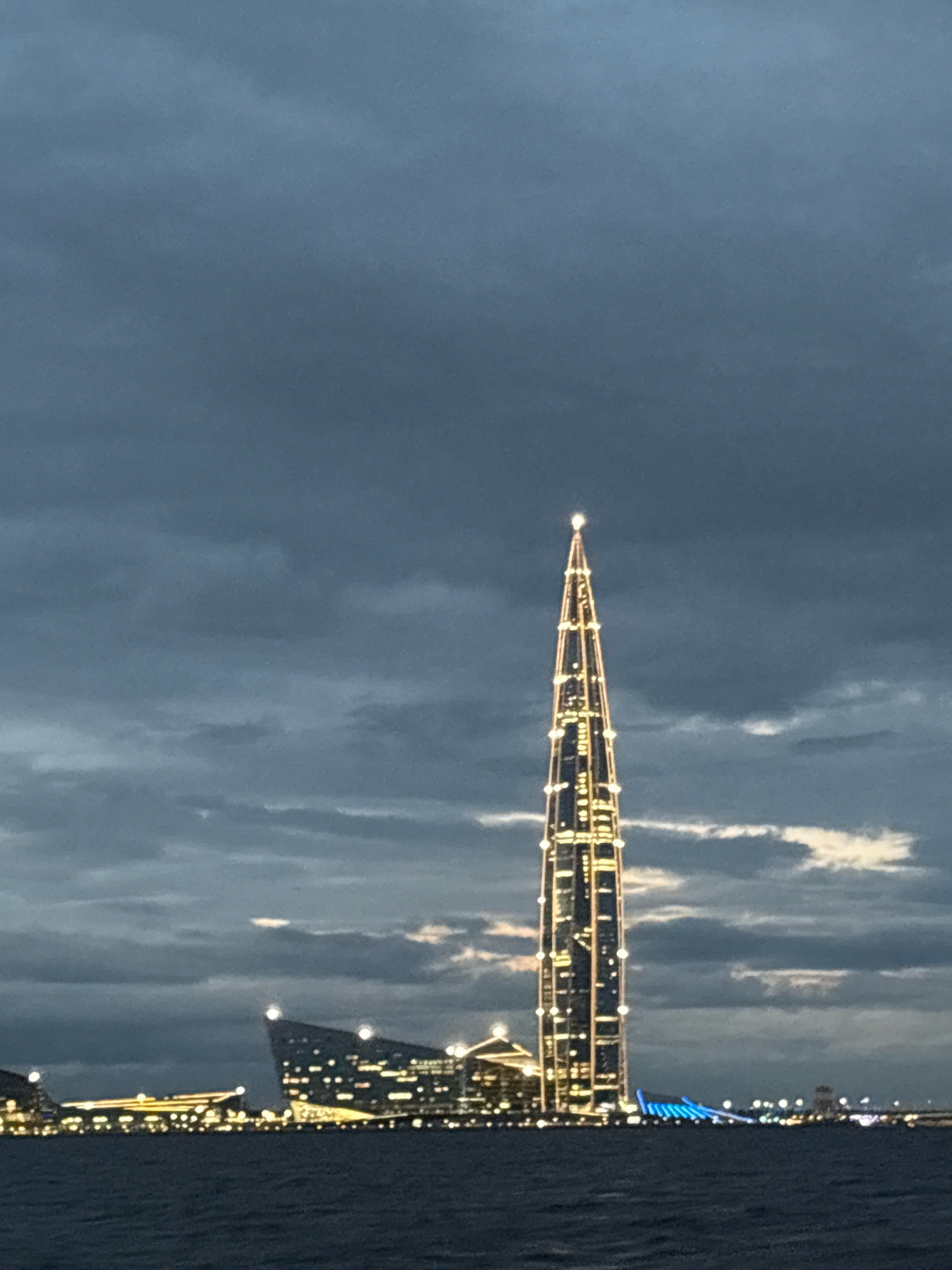 The image shows the Lakhta Center skyscraper in Saint Petersburg, Russia, at dusk. The skyscraper is illuminated with golden lights, and its pointed spire is topped with a bright light. In the foreground, there is dark blue water, likely the Gulf of Finland. In the midground, a complex of modern buildings with illuminated windows is visible, including a structure with a distinctive triangular shape. The sky is filled with dark, cloudy patterns, suggesting an overcast or stormy weather condition. The overall scene depicts a contemporary urban landscape against a dramatic sky.