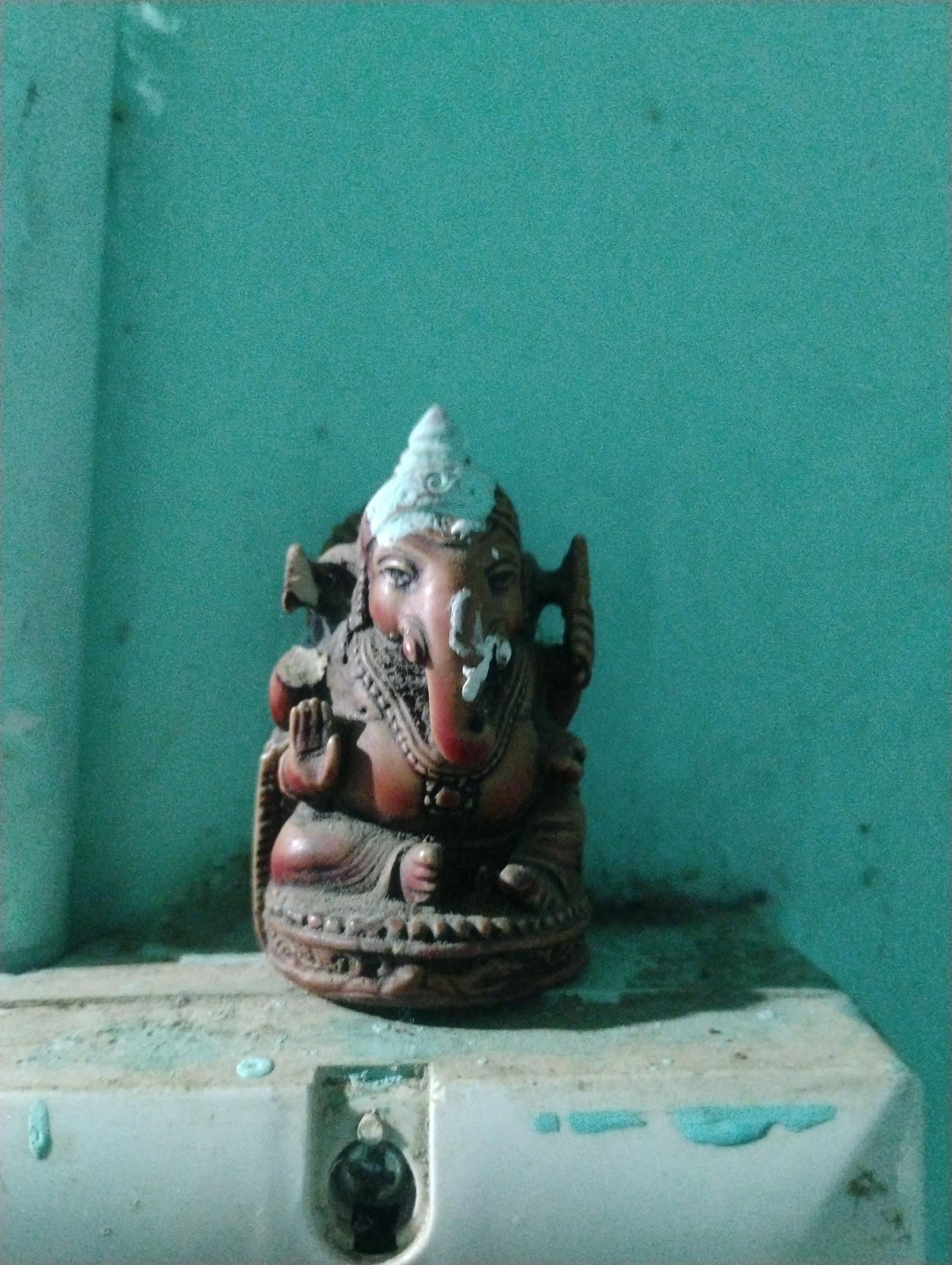 An idol of the Hindu deity Ganesha, recognizable by its elephant head and human body, is positioned centrally in the frame. The idol, appearing to be made of a reddish-brown material, is seated on a decorative base. It wears a white conical headdress and has intricate carvings on its body and base. The idol is placed on a dusty, light-colored surface, possibly a shelf or ledge, which is also marked with some light blue smudges. Behind the idol, a wall painted in a teal-green hue serves as the backdrop. The lighting in the scene is dim, casting shadows and giving the image a slightly grainy texture. The overall environment appears to be an indoor setting, with the teal wall suggesting a painted surface. No people are visible in the image.