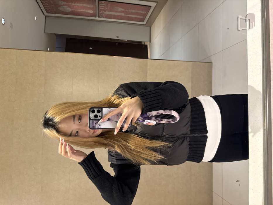 A single individual, presumed to be female, is depicted in a mirror selfie taken indoors within Yuzhong District, China. The person has long, light brown hair with visible dark roots and is wearing a black long-sleeved knitted top, a black puffer vest, a white undergarment, and black pants or leggings. Their right hand holds a dark-colored smartphone, equipped with a patterned case and a purple and white tie-dye lanyard, oriented towards the mirror. The left hand is raised, with fingers touching the hair near the crown of the head.
The background, visible through the mirror's reflection, consists of light beige tiled walls with horizontal grout lines. To the right of the individual's reflection, a hallway or room extends, featuring a dark brown door and two large, rectangular framed artworks mounted on the wall. These artworks display red backgrounds with white depictions of kitchen utensils and the partial word "KITCHEN." The floor throughout the visible area is composed of light-colored, square tiles. Recessed lighting is observable in the ceiling's reflection. A light-colored countertop is visible in the foreground, at the bottom of the image, with a faint reflection of an object in the lower right corner.