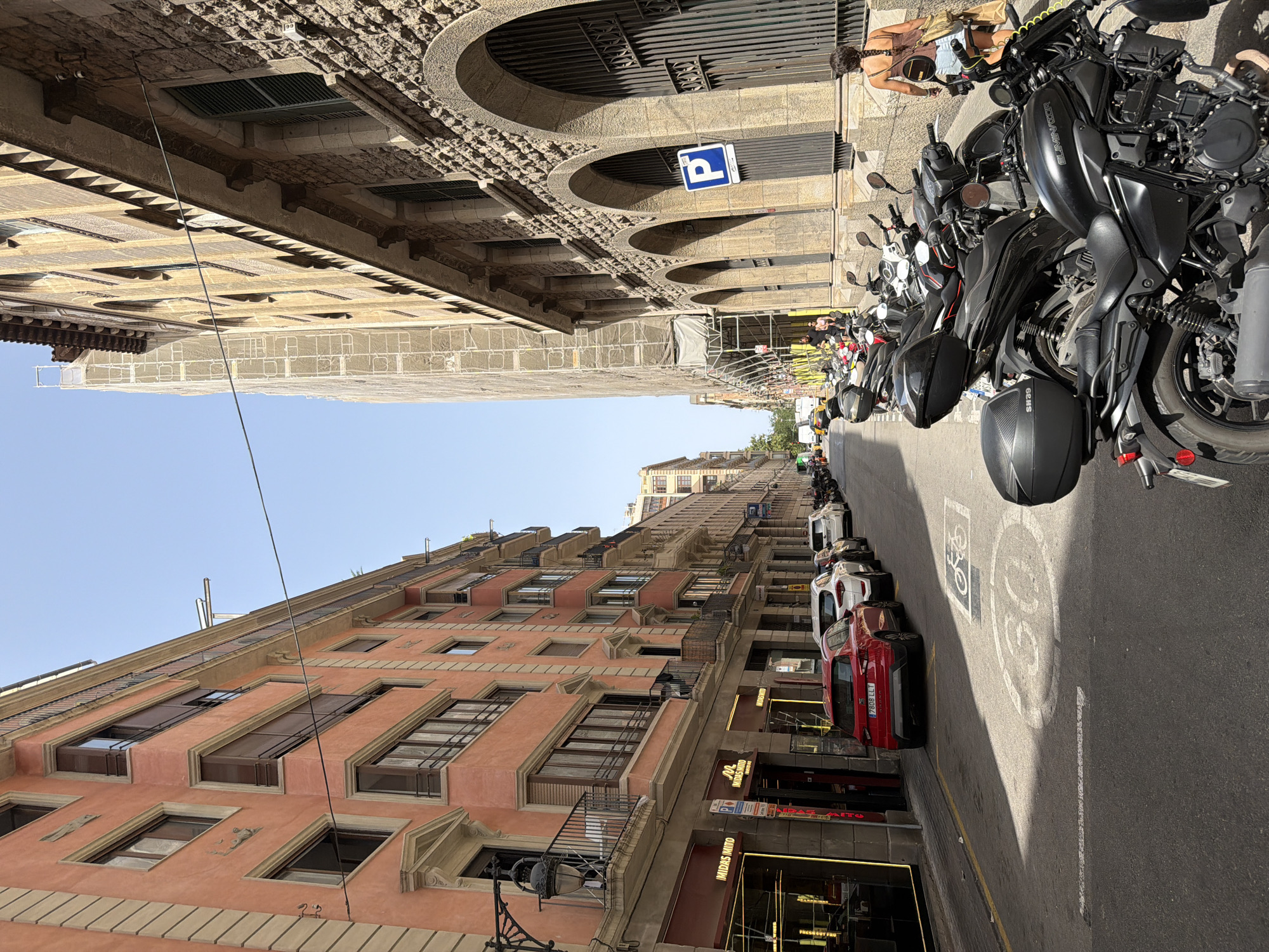 In Barcelona, Spain, a street scene is depicted with buildings lining both sides. On the left, a coral-colored building with balconies and dark-framed windows stands tall. Below, storefronts with red awnings are visible, one displaying "IMIDAS MITO". Cars, including a red hatchback and white sedans, are parked along the street. On the right, a stone building with arched entrances and barred windows is prominent. A blue parking sign is mounted on this building. A woman with braided hair walks away from the camera, carrying bags, while a person in a black shirt walks away in the distance. Numerous motorcycles and scooters are parked on the right side of the street, partially obscuring the stone building. The road surface shows painted markings, including a bicycle symbol and a "30" speed limit sign. The sky is clear and blue.