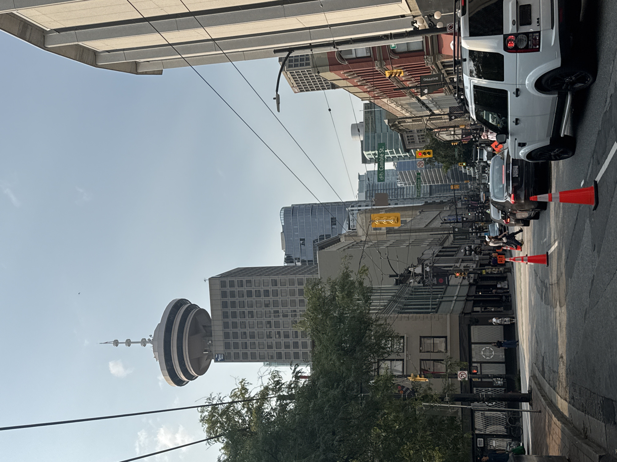 This is an eye-level, medium-shot, vertical photograph taken on a clear, sunny day, looking down a city street in Vancouver, Canada. The scene depicts an urban environment with a mix of modern and older architecture, traffic, and pedestrians.
Dominating the skyline is the distinctive observation deck of the Vancouver Lookout at Harbour Centre, a circular structure with multiple tiers atop a tall, rectangular office building. To the right of this, a lighter-colored, modern building with large vertical concrete elements stands prominently. Further down the street, a row of older brick buildings with red and cream facades line the right side, featuring arched windows on the upper floors. On the left, a lush green tree partially obscures a building with a grey facade and many windows.
The street is populated with vehicles, including a white SUV in the foreground, a dark sedan behind it, and other cars further down the road. Traffic appears to be moving slowly, with orange traffic cones placed in the street. Several pedestrians are visible on the sidewalks and crossing the street. One person is walking a dog, and others are engaged in conversations or simply moving through the urban landscape. Overhead wires crisscross the sky, common in this area of Vancouver. Street signs are visible, with one indicating "Cambie St."
The overall impression is a bustling city street scene during daylight hours, capturing the essence of urban life in Vancouver.