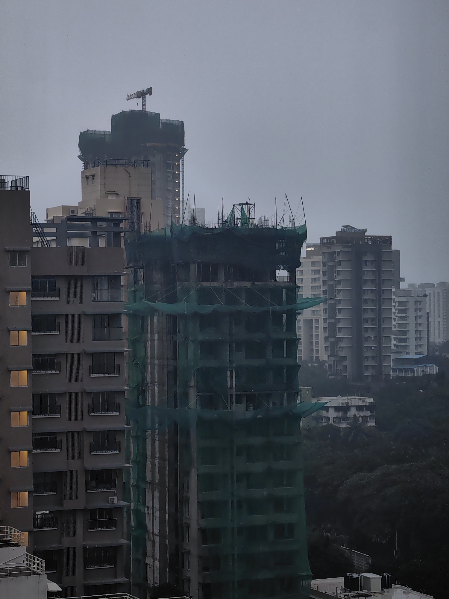 This is a medium-range shot of a city skyline on a cloudy day, likely in Mumbai, India, given the style of buildings. The scene is dominated by several multi-story buildings, some of which are clearly under construction. The most prominent structure in the foreground is a tall building covered in green netting, revealing the concrete skeleton beneath. Other buildings of varying heights and architectural styles are visible in the background, including some that appear to be residential apartments and office buildings. One building in the background has "HAPPY HOMES" written on its rooftop. A construction crane is visible atop a taller building in the distance. The sky is overcast, and the overall lighting is dim, suggesting a potentially rainy or misty day. The dense foliage of trees is visible in the lower right portion of the frame, indicating a greener area within the urban landscape. There are no people or vehicles clearly visible in the image. The focus is on the architectural elements and the ongoing construction within the urban environment.
