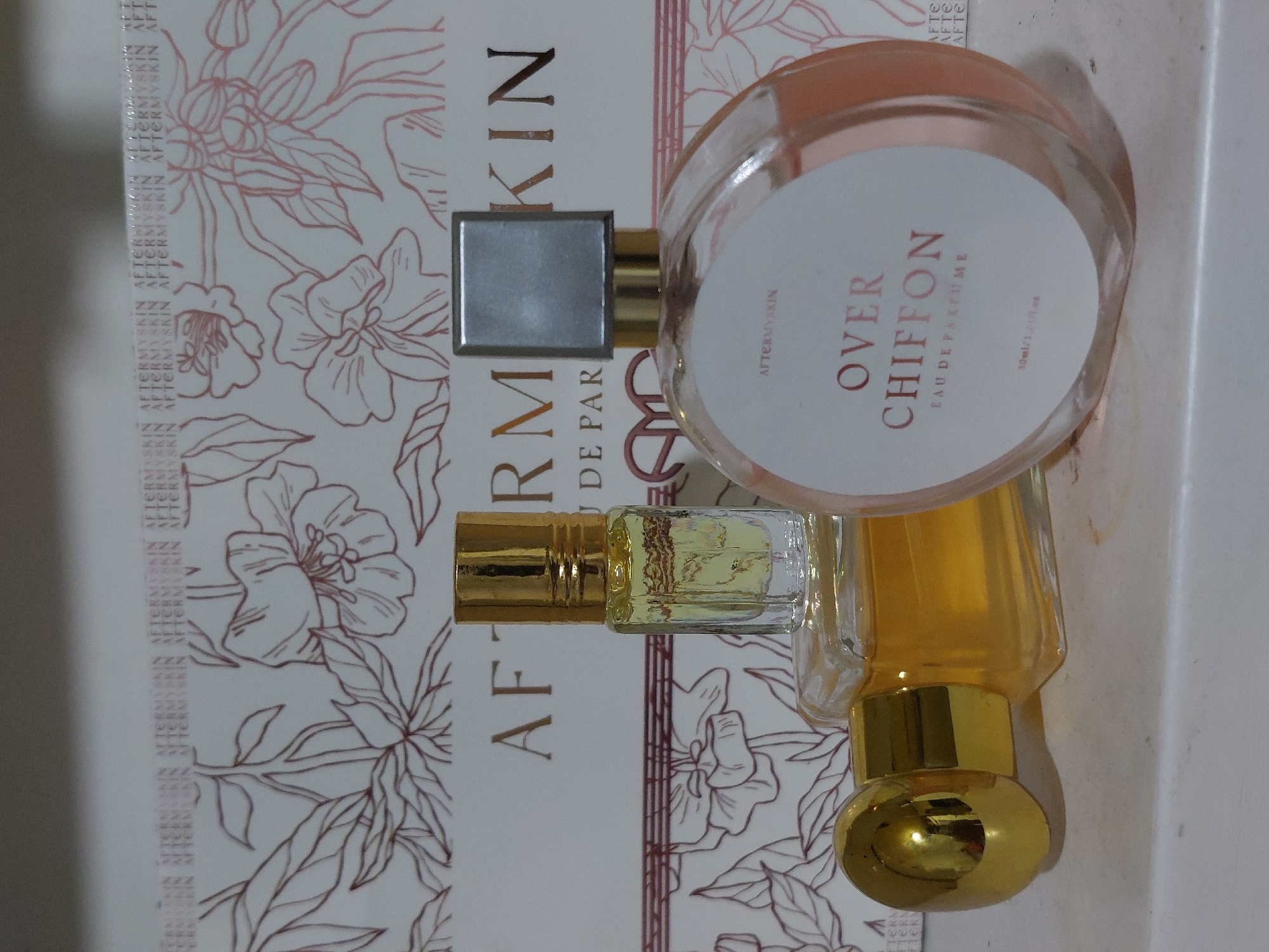 The image shows three perfume bottles. One is round and pink, with a white label that reads "OVER CHIFFON Eau de Parfum 30ml/1.0 fl.oz". The second is a small, rectangular bottle with a gold cap, and the third is a taller bottle with a gold cap and a bulbous applicator. The bottles are placed on top of a decorative box with a floral pattern and the repeating word "AFTERMYSKIN". The box's design features a color scheme of white and a muted reddish-brown. The image does not provide any information about the location or city of Lhokseumawe, Indonesia, or any people or interactions.