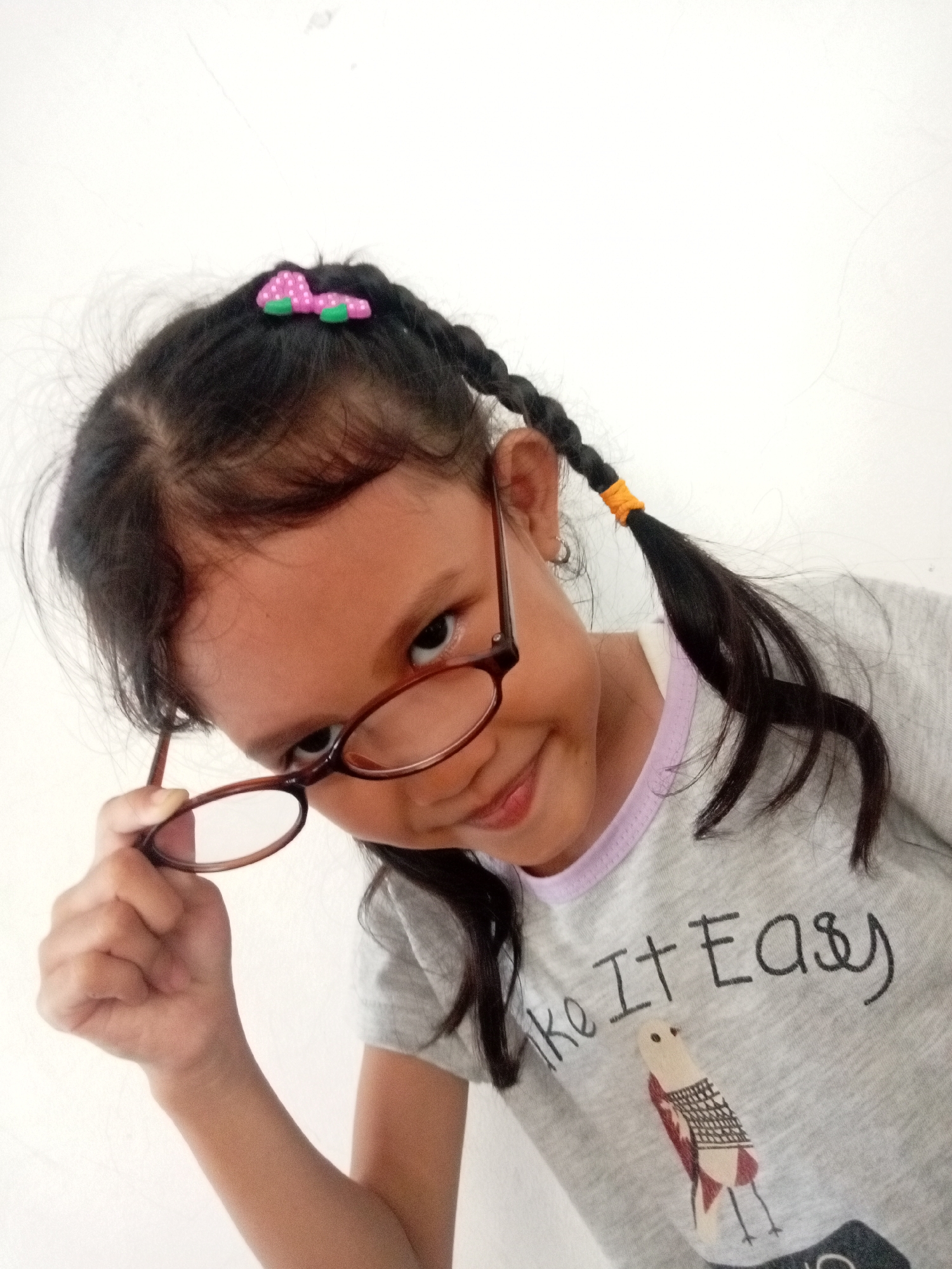 A young girl with braided pigtails and a pink bow is looking at the camera from a low angle, holding her glasses up to her chin with her right hand. She is wearing a grey t-shirt with the text "ke It Easy" and a cartoon bird printed on it. The background is a plain white wall. This image was taken indoors, likely in a residential setting in Batam, Indonesia.