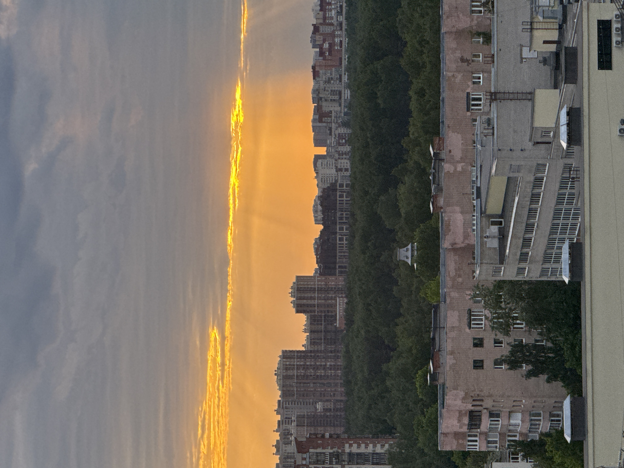 The image presents a dusk urban landscape, likely of Unknown, Russia. Dominating the foreground is a cityscape with a line of trees separating the buildings from the sky. The sky displays a vivid sunset with streaks of orange and yellow light breaking through clouds. Various buildings of different heights and architectural styles populate the scene. The buildings vary in color, from pale grey to brick red. A large building is located on the right, with multiple windows and ventilation units. The overall scene exudes a sense of urban life against a backdrop of natural beauty.