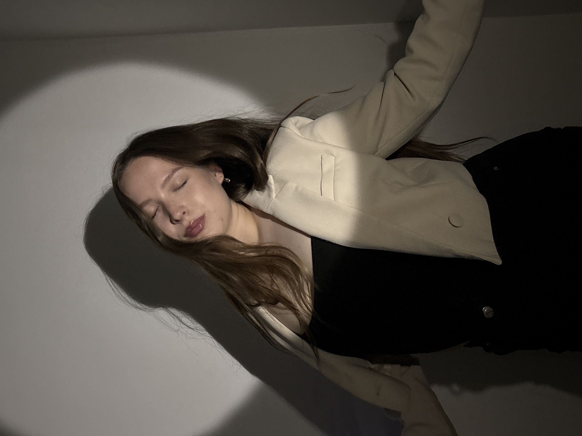 A young female, with long brown hair, is positioned in what appears to be an indoor setting against a light-colored, unadorned wall. Her eyes are closed, and her head is tilted slightly upward and to her right. Her lips are slightly parted, exhibiting a glossy texture. She wears a black strapless top beneath an open, cropped, light beige or cream-colored blazer. The blazer features notched lapels and two visible buttons on the left front panel. A small silver-toned earring is visible in her right ear. Her arms are extended slightly outwards, and a portion of black pants or skirt with a silver rivet is visible at her waist. The scene is illuminated by a direct, circular light source, casting a bright, defined circle on the wall behind and to the upper left of the subject. This lighting creates a distinct, elongated shadow of her head and upper body on the wall to the left, with additional softer shadows present on the right side of the frame. The immediate environment is featureless beyond the wall and the subject. There is no discernible information in the image to indicate a specific geographic location such as the city of Chaikovsky, Russia.