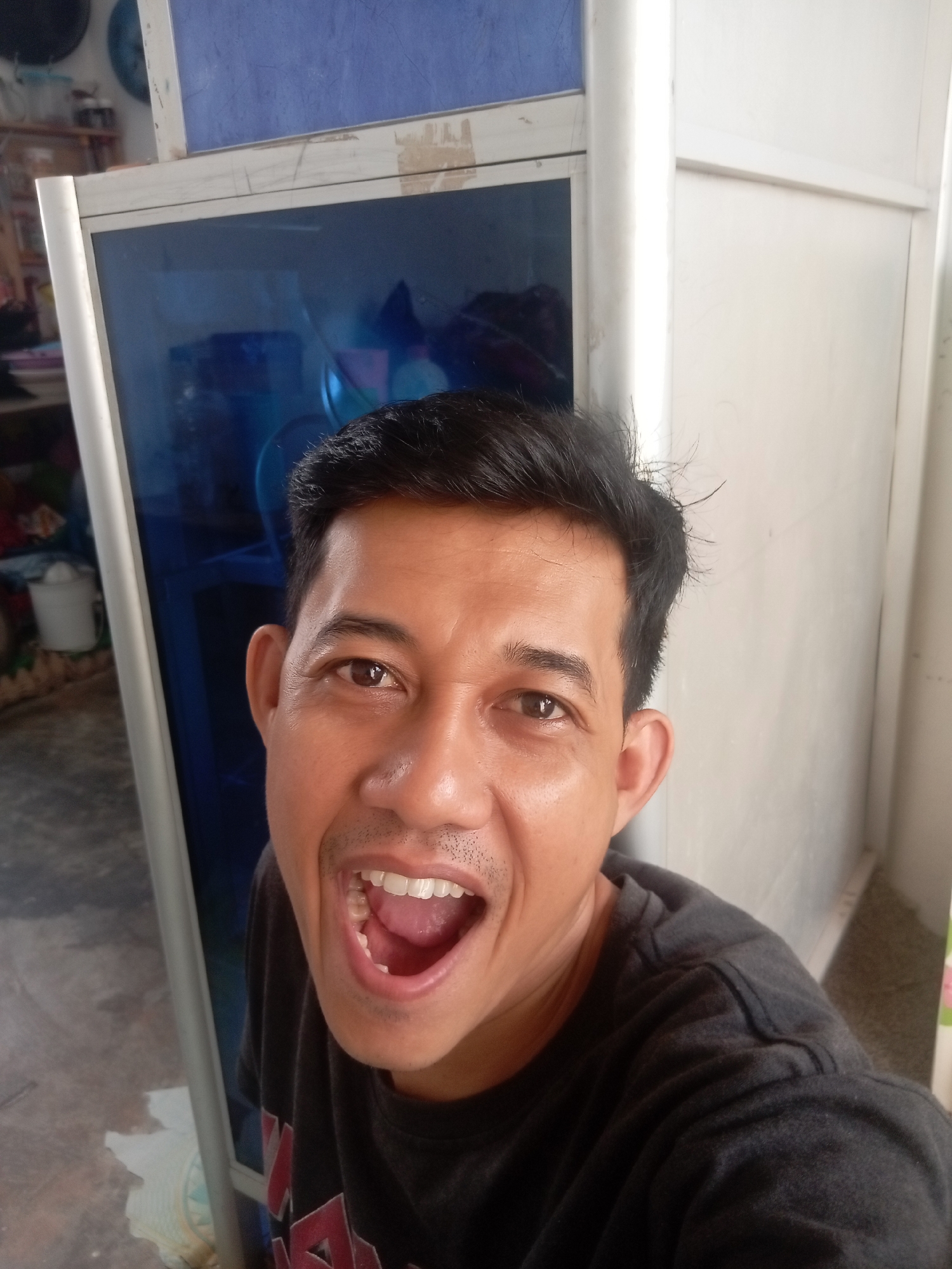 A young Asian man is taking a selfie in what appears to be a modest indoor setting. He is positioned in the center of the frame, looking directly at the camera with his mouth wide open in a surprised or excited expression. His hair is dark and short, with some strands appearing slightly disheveled. He is wearing a dark, possibly charcoal gray, t-shirt.
Behind him, to the left, is a tall, narrow cabinet with a dark blue mirrored or tinted glass front. The cabinet is framed by a silver-colored metal edge. To the right of the man, a plain, light-colored wall is visible, possibly made of painted panels or plaster. The floor in the foreground is a mottled gray concrete. In the background, on the left, a shelf with various items is partially visible, including what looks like a white bucket. The overall environment suggests a residential or small commercial space, perhaps a storage room or a less-decorated part of a home.
Without further information, it is not possible to pinpoint the specific location within Batam, Indonesia. However, the scene depicts a casual and informal moment captured by the individual himself.