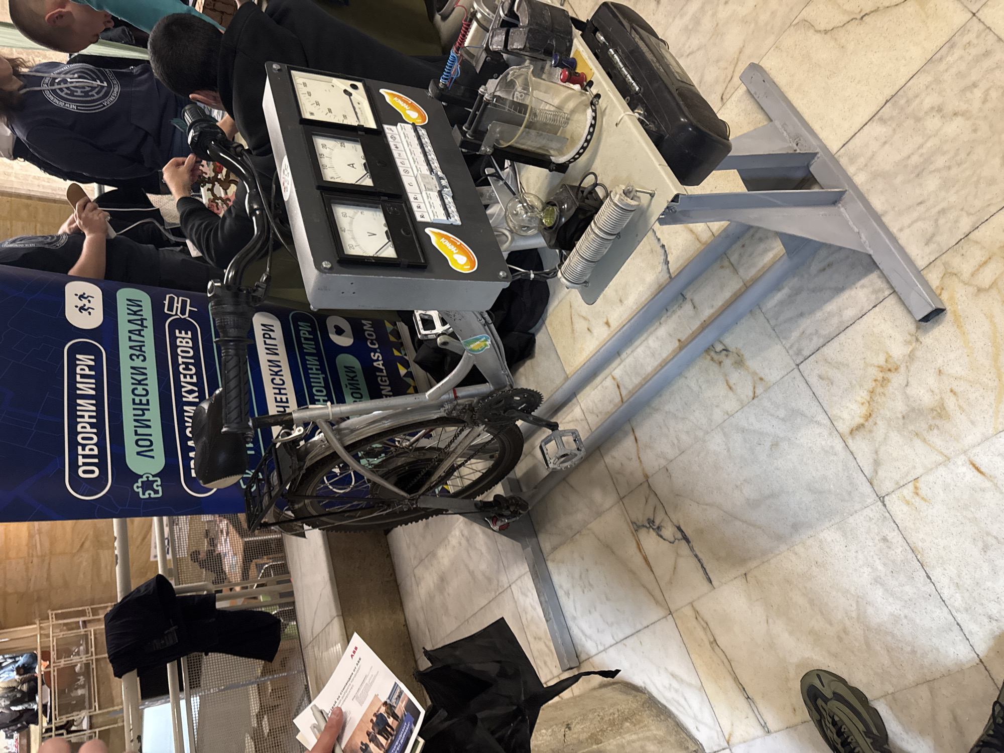 The image shows a stationary bicycle attached to a device with analog meters and electrical components, possibly for demonstrating energy generation. The bicycle is positioned on a grey metal frame, elevating it from the tiled floor. There are people around the device, appearing to observe or interact with it. A blue banner with white text in Bulgarian promoting team games and logic puzzles can be seen next to the bicycle. In the background, there are architectural features like columns and a staircase suggesting the scene might be indoors in a public space. A person in the bottom right corner is wearing sneakers and standing near the device. Another person is holding a brochure or document with an "ABB" logo. The location is Plovdiv, Bulgaria.