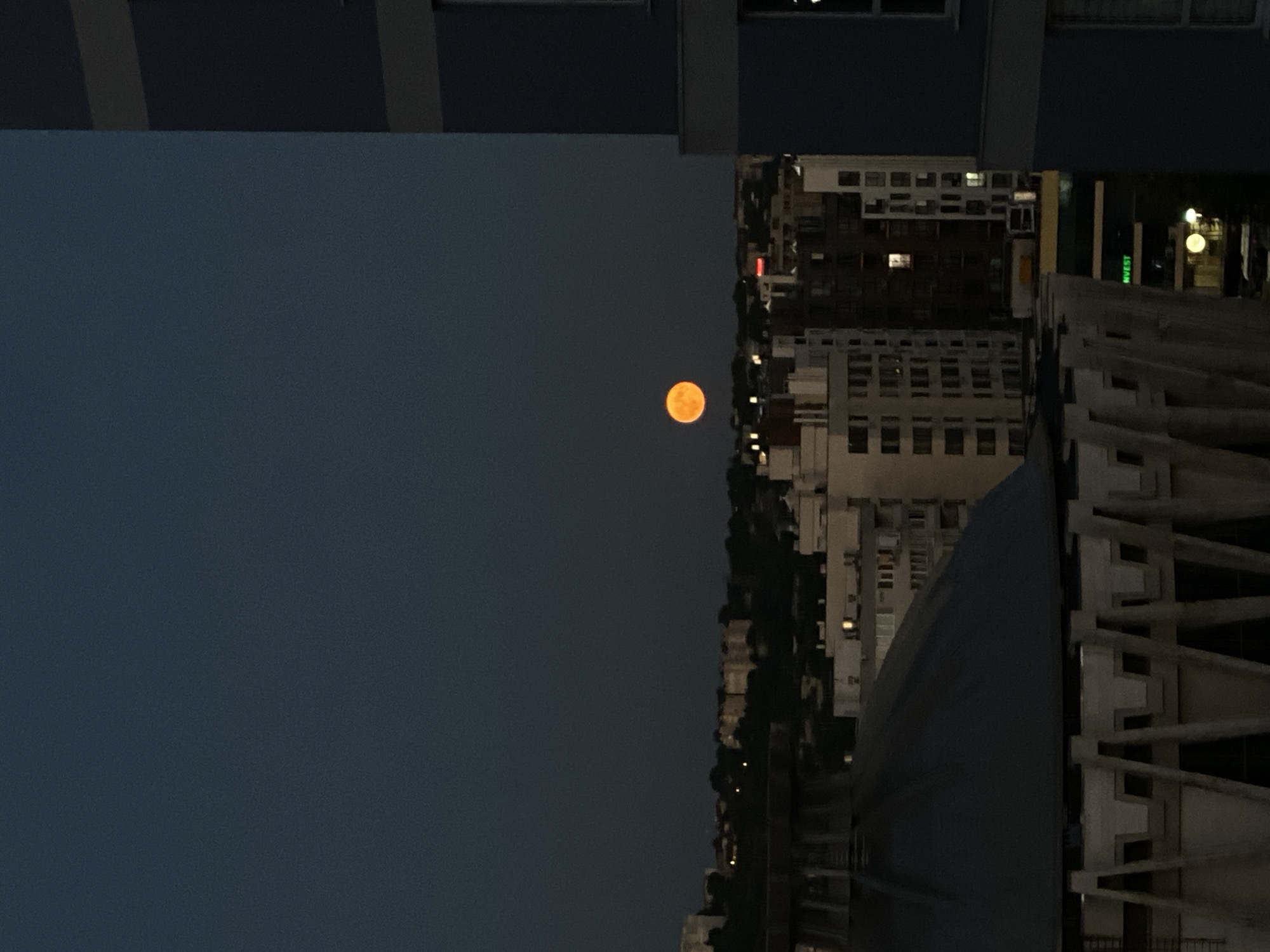An orange moon hangs in a dark blue twilight sky over a city skyline. The moon is located in the upper center of the image. In the foreground, a large, dark, conical structure with a ribbed underside dominates the lower left. To the right, a modern blue building rises vertically, partially obscuring the view of the city. The city itself is a dense collection of buildings of varying heights and architectural styles, with lights visible in some windows. The overall impression is of an urban landscape at dusk.