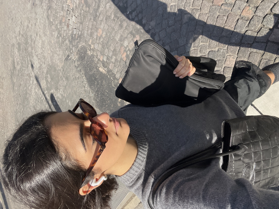 A single individual, an adult female, is depicted from an elevated perspective, walking on a Parisian street or sidewalk paved with greyish-brown cobblestones. The scene is under bright daylight, casting distinct, elongated shadows, including one resembling a street lamp. The female has short, dark hair and wears tortoise-shell oval sunglasses with reddish lenses, along with a white wireless earbud in her left ear. She is dressed in a grey crew-neck sweater and dark lower garments. On her left shoulder, she carries a black shoulder bag with a textured, embossed pattern and a small, unidentifiable logo patch. In her right hand, she holds a black padded laptop sleeve or portfolio featuring a zipper and white piping. Her right hand displays manicured nails. The individual appears to be in motion, looking slightly downward and to her right.