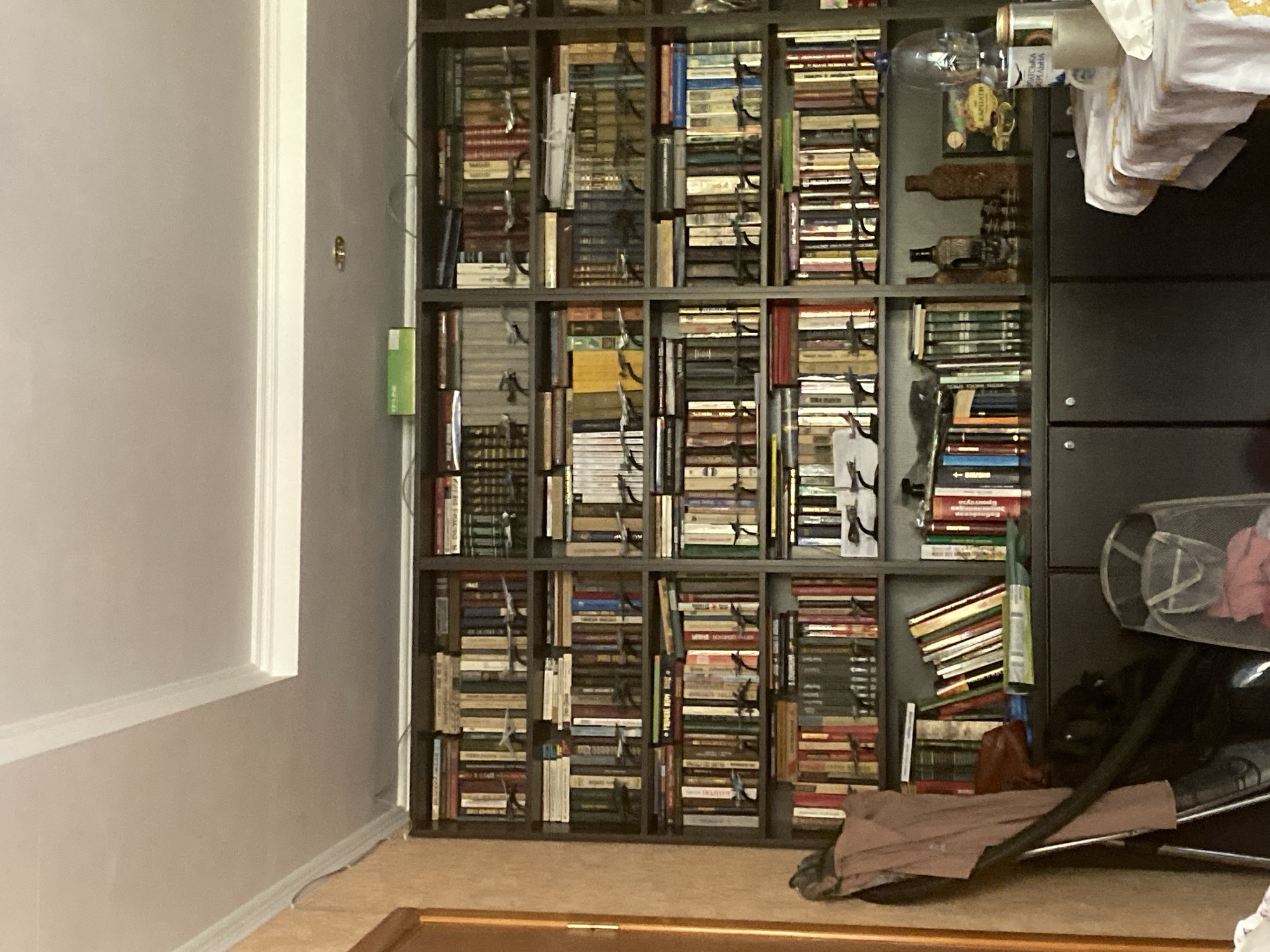 The image shows a tall, dark wood bookshelf filled with numerous books. The books are arranged in vertical rows, with some shelves also containing small model airplanes. The bookshelf is divided into multiple sections by vertical and horizontal dividers. On top of the bookshelf, there are a few items, including a green box and some wires. Below the bookshelf, there is a dark cabinet with several doors. To the left of the bookshelf, a portion of a wooden door frame is visible, and a tan-colored wall is to its left. A brown garment is hanging on the left side of the bookshelf. To the right of the bookshelf, a table covered with a white tablecloth featuring a checkered pattern is visible. On the table, there are several objects, including a clear plastic bottle, a mug, and a can. The ceiling above the bookshelf is white and has decorative molding.