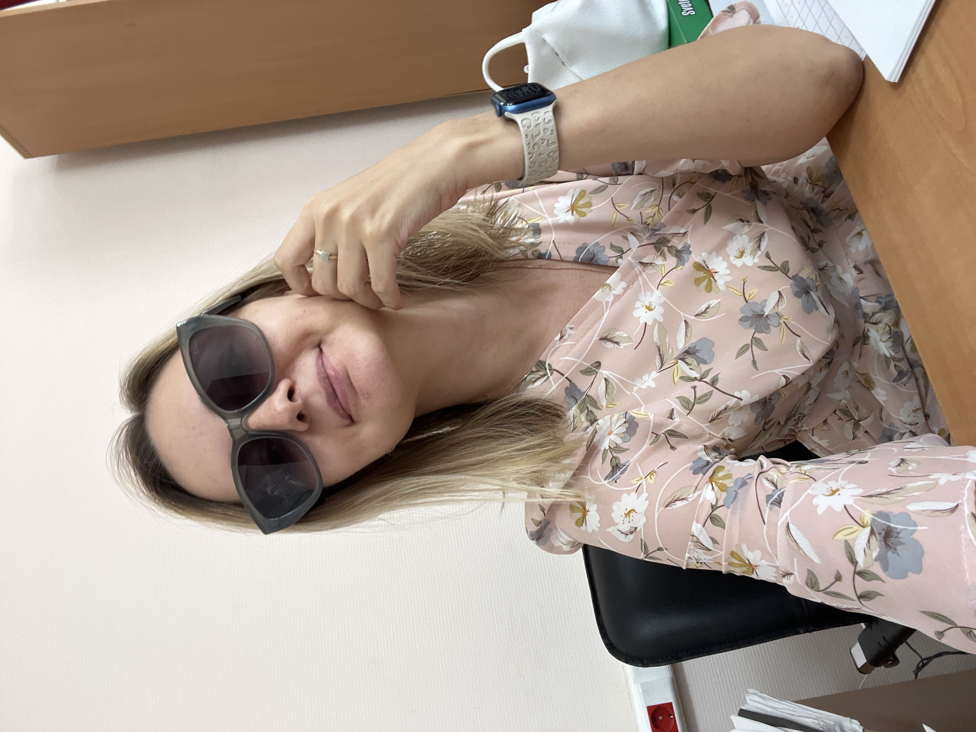 A woman with blonde hair and sunglasses is sitting at a desk in an office. She is wearing a pink floral dress and a watch on her left wrist. A white bag is visible behind her on the right. The scene appears to be a typical office environment. There is no specific information in the image to indicate the location is in Volgograd, Russia.