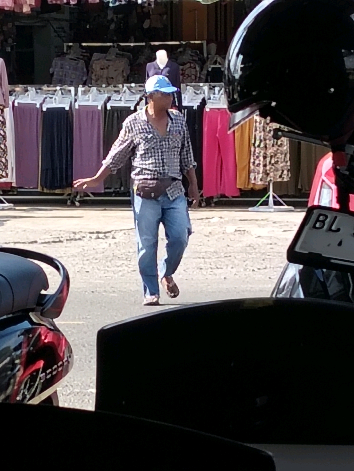 A man in a plaid shirt, blue cap, and jeans walks across a street in front of a market stall in Grong-Grong, Indonesia. He is wearing a brown fanny pack and sandals. The market stall displays various colorful clothing items. A black helmet and a motorcycle license plate are visible in the foreground.