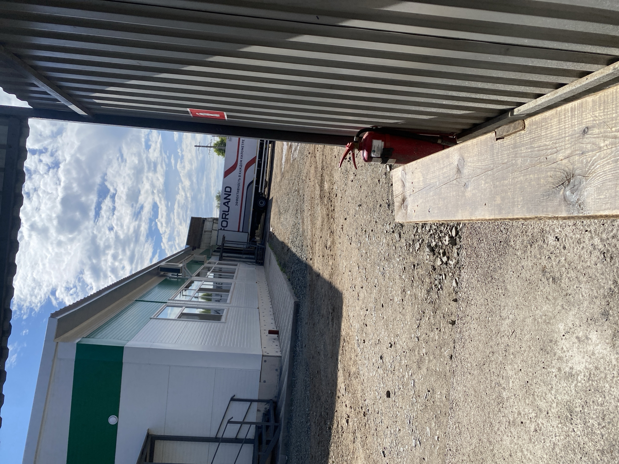 The image depicts an outdoor scene on a sunny day with a partly cloudy sky. In the foreground, a weathered wooden plank rests on the ground, partially obscuring a red fire extinguisher leaning against a corrugated metal wall. The wall appears to be part of a building or structure.
To the left of the metal wall and wooden plank, a white building with green trim is visible. The building has windows and a ramp leading up to an entrance with a metal railing. In the background, a white truck with the word "FORLAND" and Russian text ("ЭФФЕКТИВНОСТЬ В КАЖДОМ КИЛОМЕТРЕ" which translates to "Efficiency in every kilometer") is parked. The ground is unpaved, covered with dirt, gravel, and some scattered debris.
The scene appears to be an industrial or commercial area, possibly a logistics hub or a construction site. The presence of the fire extinguisher suggests safety measures are in place. The overall impression is one of a functional, utilitarian environment. There are no people visible in the image. The location is identified as being in the city of Orenburg, Russia, based on the provided information.