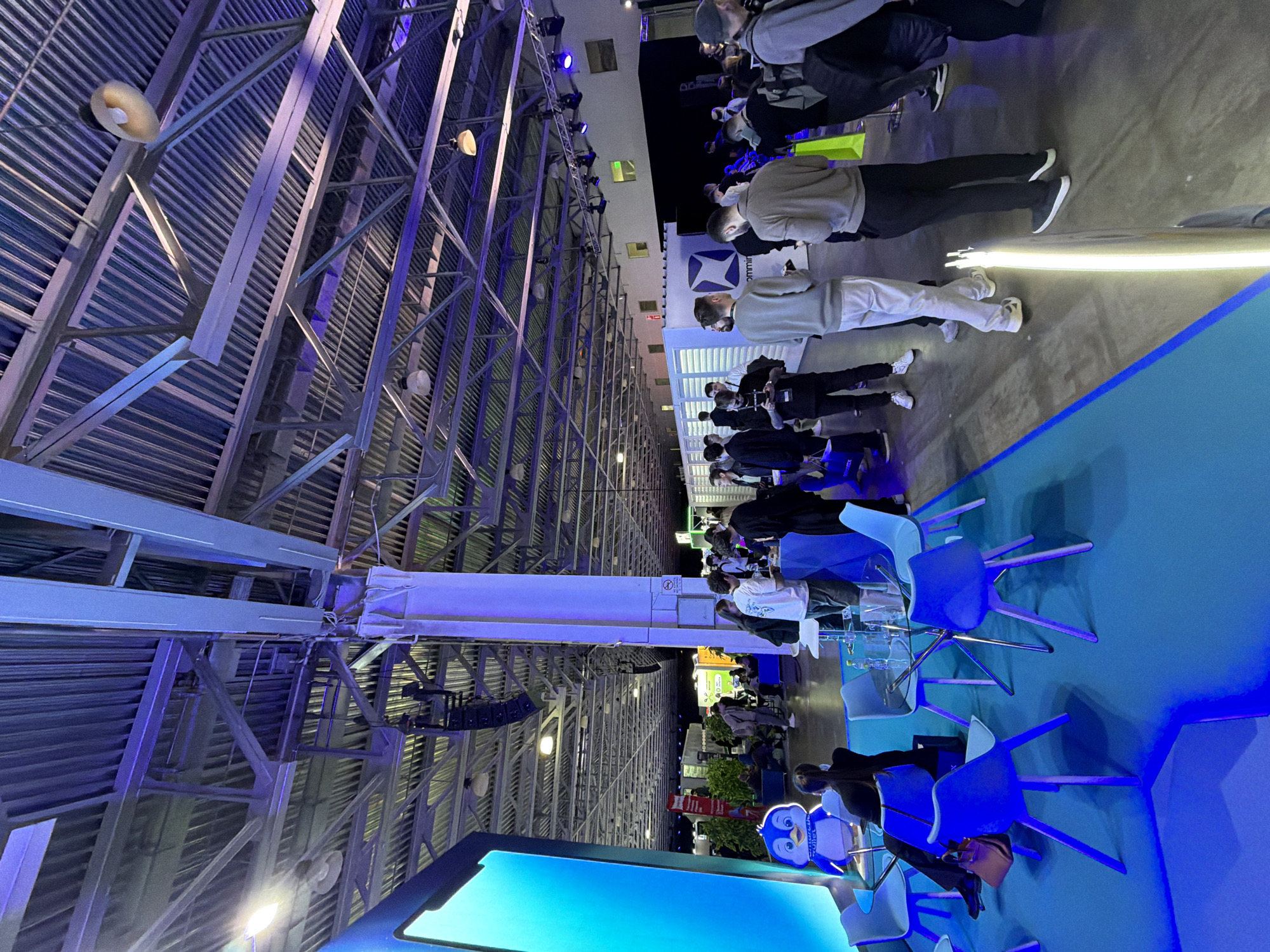 The image shows the interior of an event space, likely a convention center or exhibition hall in Khimki, Russia. The scene is oriented in a vertical fashion. The industrial ceiling with a metal support structure is visible. Groups of people are standing and walking, with some wearing face masks. Blue lighting is used, creating a modern ambiance. There are rows of blue chairs and a large screen displaying a blue background. The room is populated with various exhibition stands and displays. The overall scene appears to be a professional event or gathering.