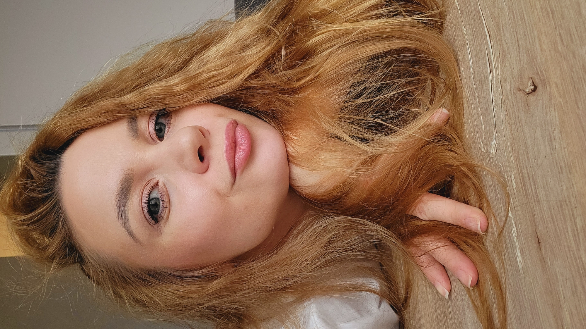 A portrait of a young woman with long, wavy, reddish-blonde hair is oriented sideways. Her face is fair-skinned with light makeup highlighting her eyes and lips. She is lying on a light brown wooden surface, possibly a tabletop. Part of her hand is visible in the right side of the frame, resting on the wood alongside her hair. A small detail, potentially a knot or imperfection in the wood, is present near the top-right corner of the image. The background is out of focus. There is no specific indication of the location being Chaikovsky, Russia, as the setting appears to be indoors and generic.