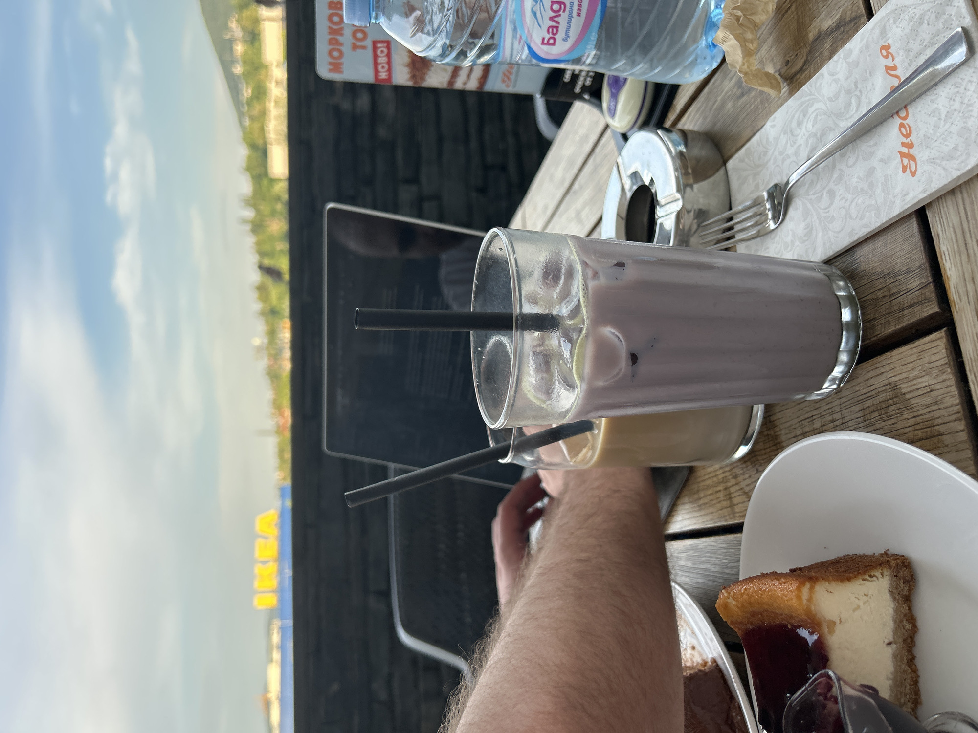 In the city of Sofia, Bulgaria, a table at an outdoor cafe displays a cheesecake with berry compote, a glass of creamy beverage with ice and blueberries, a glass of coffee with cream, a laptop, an ashtray with a cigarette butt, a bottle of mineral water, a fork, and a napkin with Cyrillic script. In the background, the IKEA logo is visible, suggesting the cafe is located in proximity to an IKEA store, likely on the outskirts of Sofia. The weather appears to be overcast with a hint of sunlight breaking through.