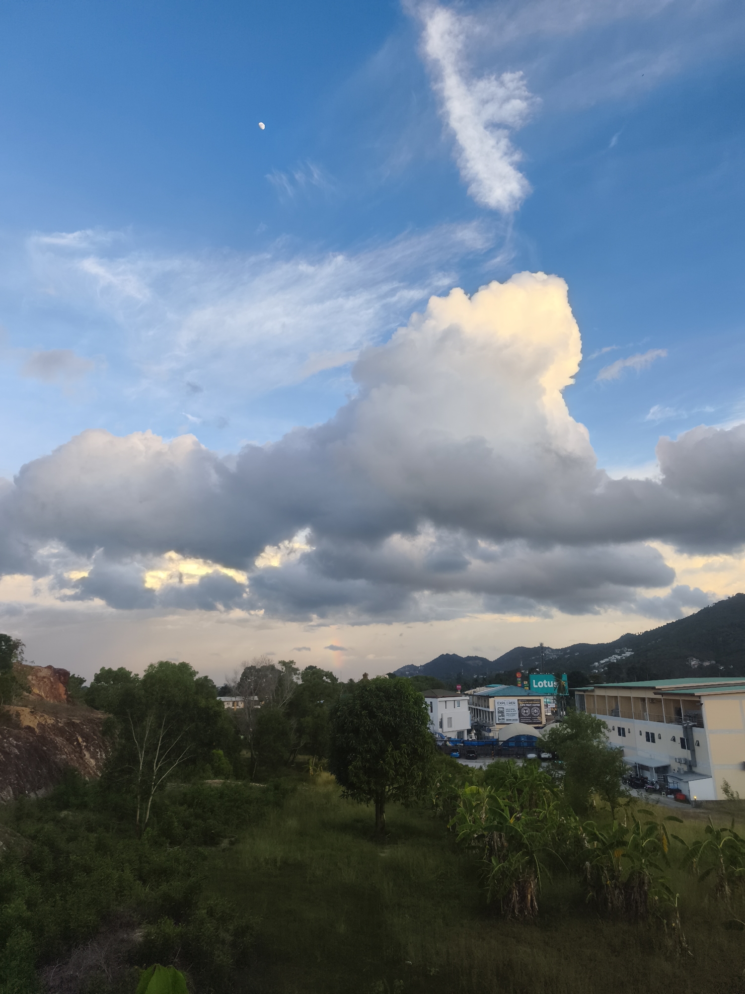 The image captures a picturesque landscape scene in the city of Ko Samui District, Thailand. The sky is a vibrant blue, adorned with a crescent moon and a variety of clouds, ranging from wispy cirrus formations to large, cumulus clouds. Some of the clouds are illuminated by the sun, displaying a warm, golden hue, while others appear darker, suggesting an approaching storm or the setting sun.
In the mid-ground, a lush green landscape unfolds, characterized by dense vegetation and trees. A rocky outcrop with a reddish-brown hue is visible on the left side of the image, adding a touch of natural ruggedness to the scene. Beyond the greenery, a cluster of buildings and structures are spread across the landscape, some of which are brightly lit. A prominent billboard with the word "Lotus" in green lettering is visible, along with other signs indicating commercial establishments. The buildings appear to be a mix of modern architecture, with some featuring light-colored facades and balconies. Hilly terrain covered in trees forms the backdrop of the scene. The overall impression is one of a serene and scenic location, possibly a residential or commercial area nestled within a natural environment.