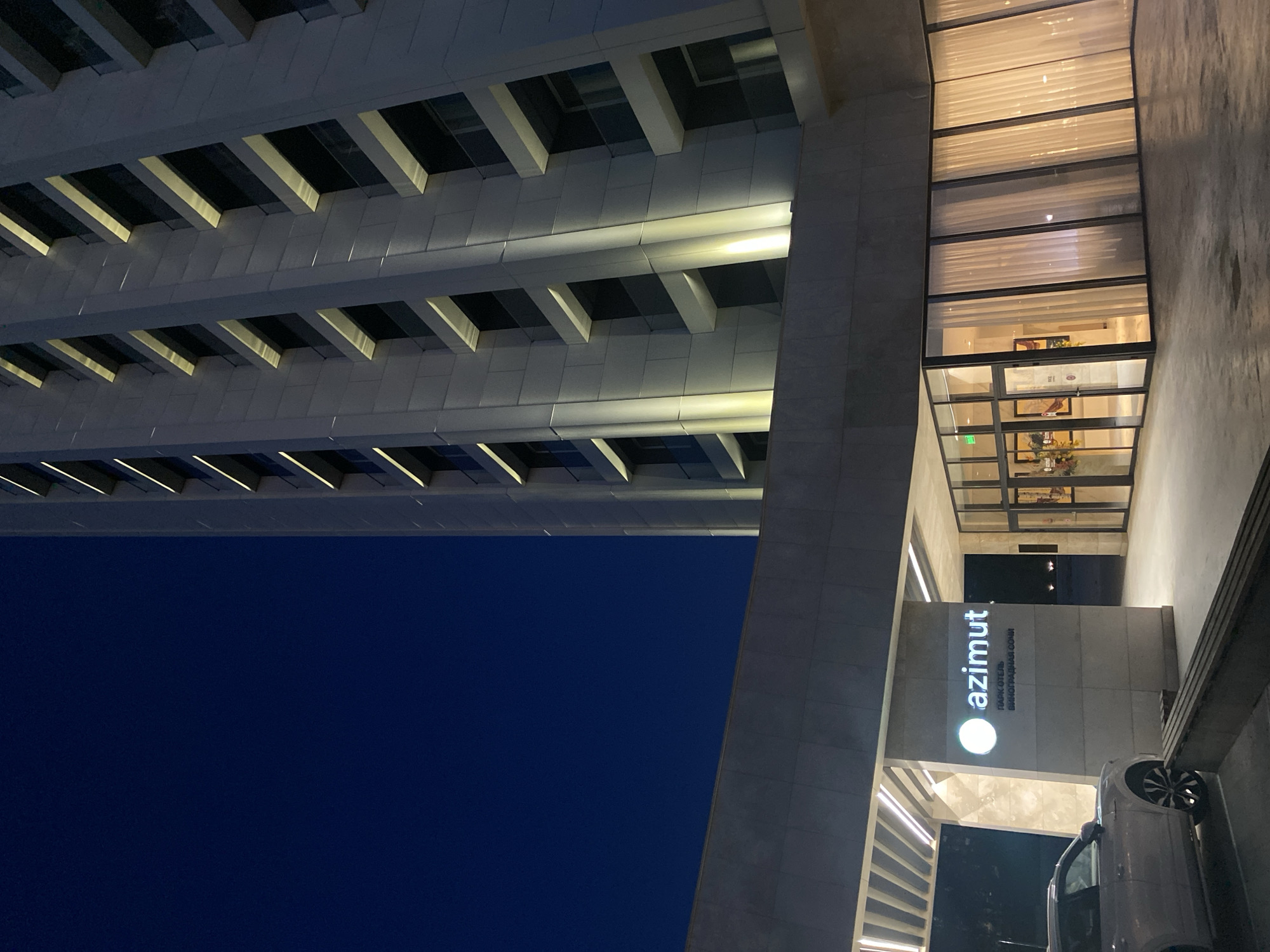 The image shows an exterior view of the Azimut Hotel in Sochi, Russia. The building has a modern architectural design, featuring prominent horizontal ledges with recessed lighting beneath each one. To the right of the building, there is a glass-enclosed section that provides a view into the hotel's interior, including furniture and decorations. The "azimut" hotel sign is prominently displayed near the entrance. A car is partially visible near the entrance. The sky is dark, indicating that the photo was taken during the evening or night.