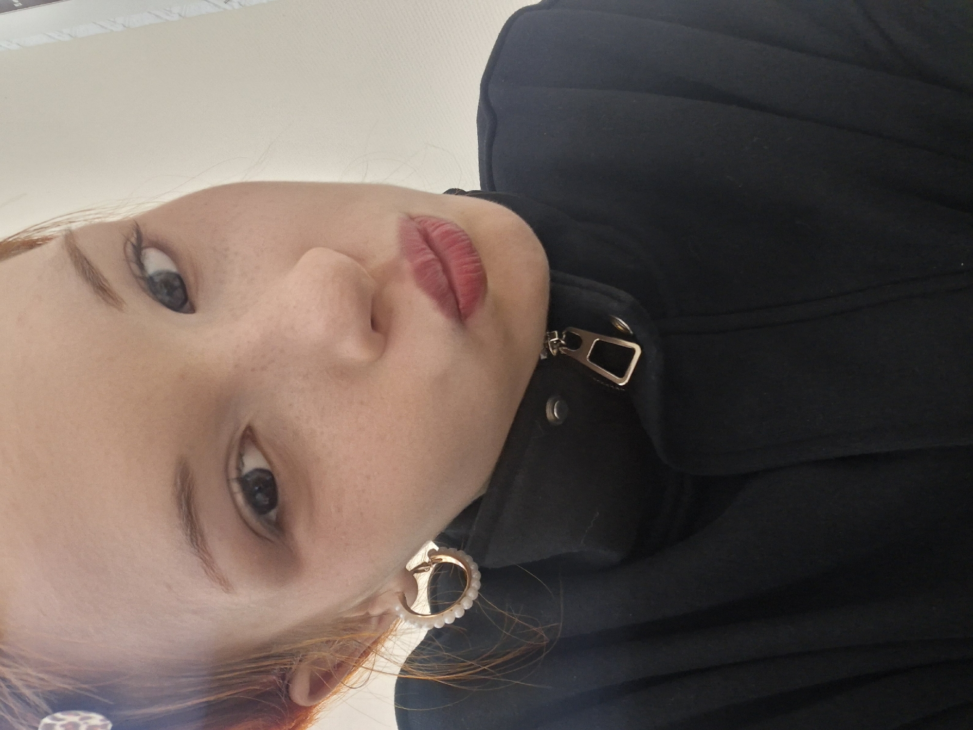 A close-up, rotated portrait features a young person with fair skin and visible freckles. The subject has reddish-brown hair, light-colored eyes (appearing blue or grey), and is wearing a bold red or berry-toned lipstick. Their expression is neutral, and their gaze is directed upwards towards the camera.
The person is dressed in a dark, likely black, jacket or coat, which features a prominent gold-colored zipper pull and a metallic rectangular buckle or loop near the collar area. On their visible right ear, a gold-toned hoop earring adorned with small white beads or pearls can be seen. The background is a plain, light-colored surface, suggesting an indoor setting. There are no discernible clues regarding the time of day or weather. No text is visible in the media file. The provided context indicates the location is Chaikovsky, Russia.