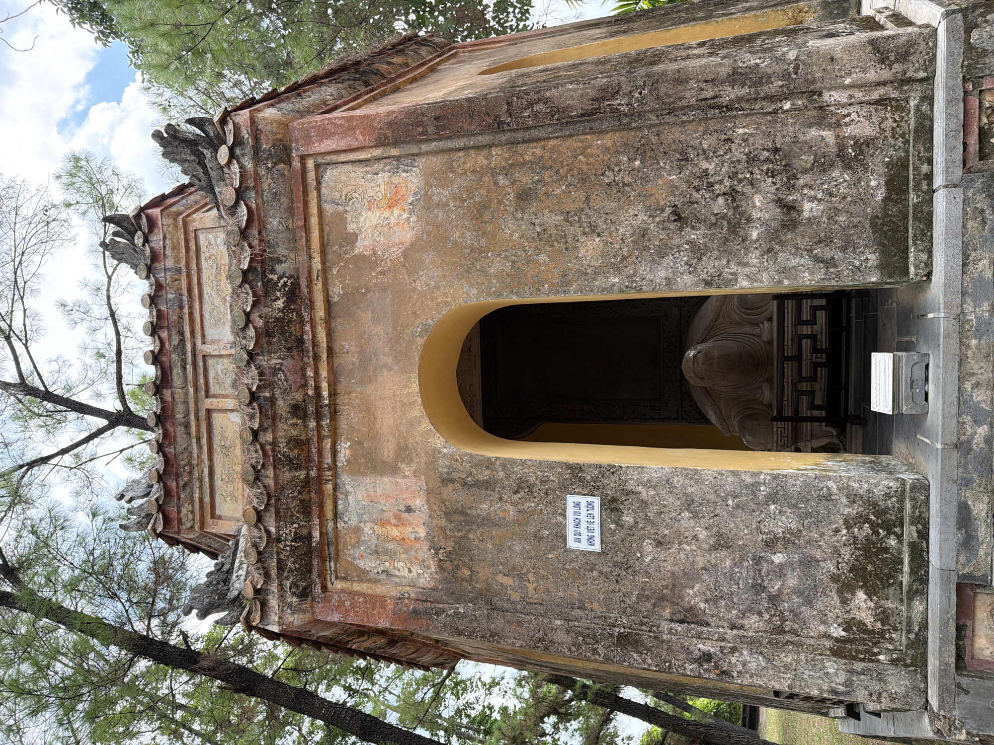 This is a low-angle, full shot of an ancient, weathered tomb in Kim Long Ward, Vietnam, possibly part of a historical cemetery or imperial tomb complex. The structure is octagonal, with a multi-tiered, tiled roof featuring decorative dragon motifs. The walls are made of stone and have a distinct patina of age, with patches of moss and lichen visible.
An arched entryway in the front of the tomb leads to a darker interior. Inside, a large, sculpted stone turtle is partially visible, positioned on a dark, carved wooden platform. To the left of the entryway, a white sign with Vietnamese text is affixed to the wall. The tomb is surrounded by lush greenery, with tall trees and their branches extending over and around the structure. The sky above is a mix of blue and white clouds, with dappled sunlight filtering through the trees. The foreground shows a stone base and steps leading up to the tomb, also showing signs of age and wear.