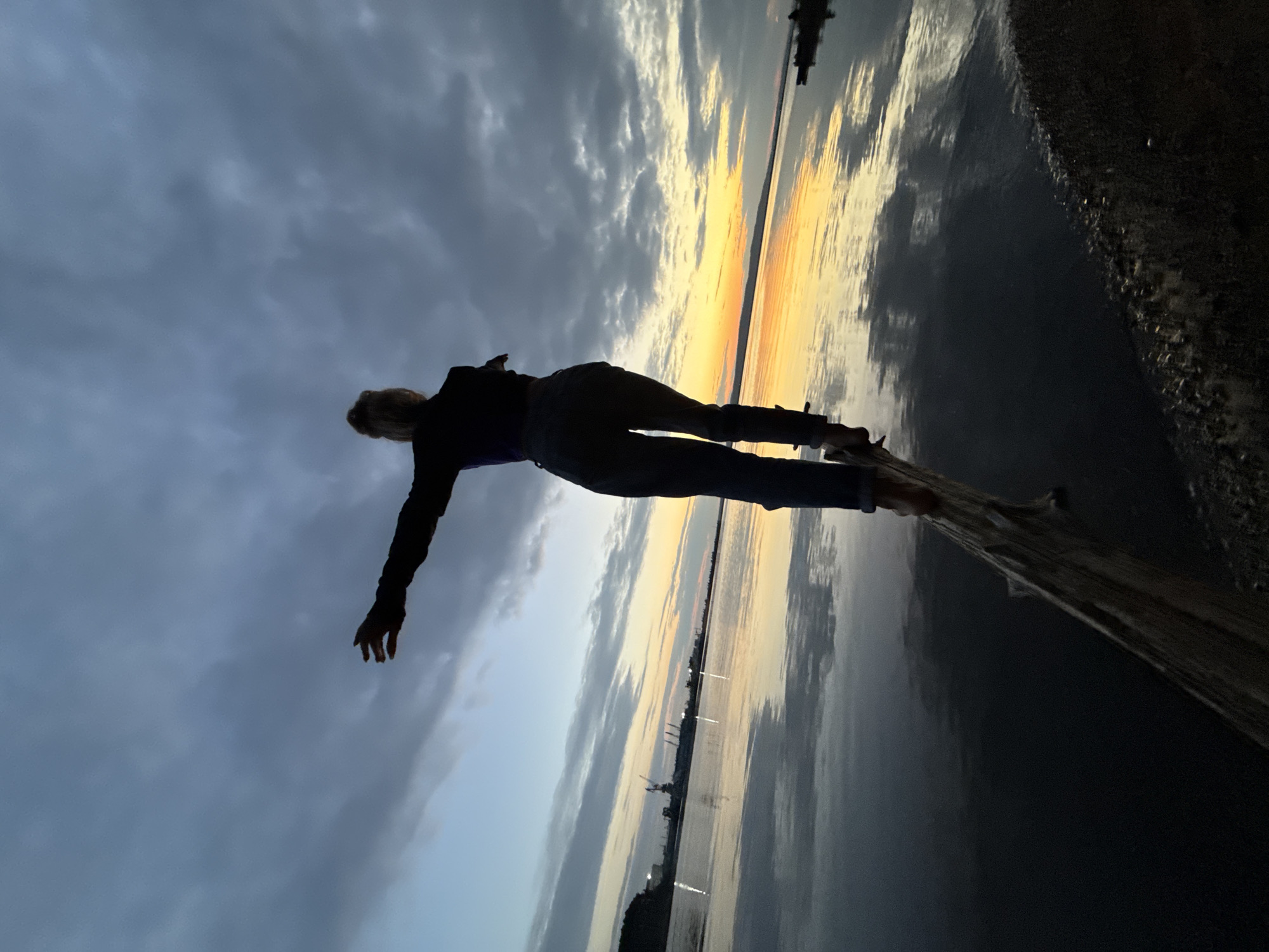 A person, silhouetted against a twilight sky, stands with arms outstretched on a fallen tree trunk at the edge of a body of water. The sky is filled with dramatic clouds, with a band of orange and yellow light visible on the horizon, suggesting a sunset. The water reflects the sky and the distant lights of what appears to be an industrial area with cranes and buildings on the far bank, likely within the city of Chaikovsky, Russia, given the proximity to the Kama River reservoir. The person is wearing dark clothing, including jeans and what looks like a jacket, and is balancing on the log with their feet. The scene conveys a sense of peaceful solitude and contemplation amidst the natural beauty of the fading light and the urban industrial landscape.