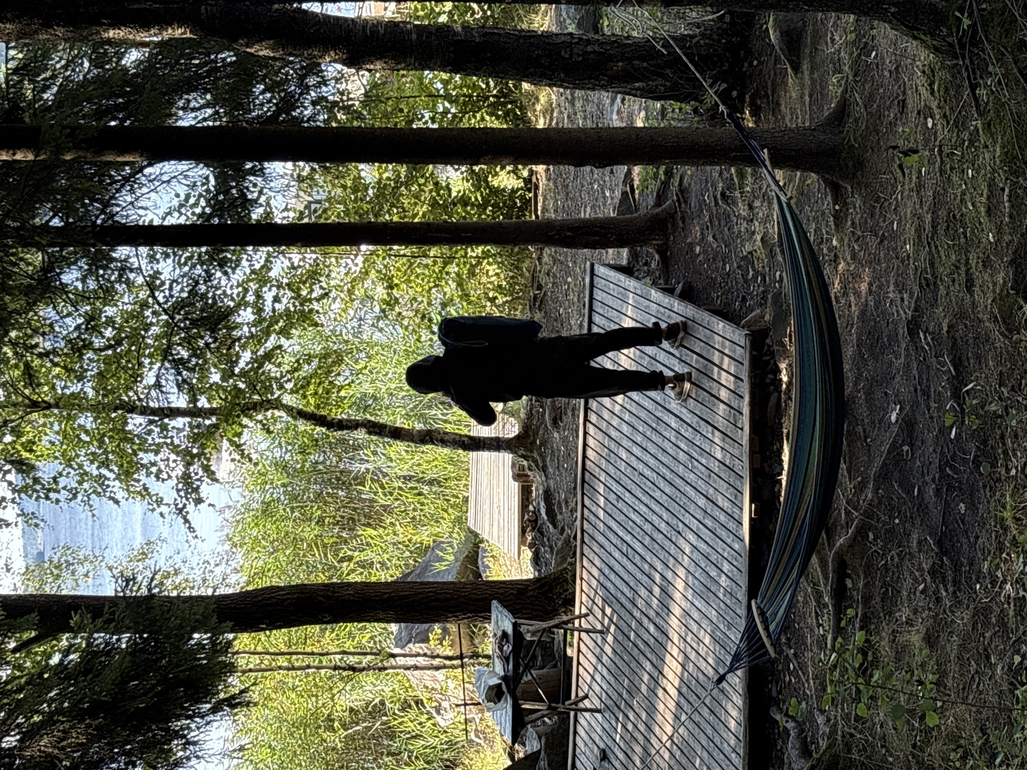 The image depicts an outdoor scene at a lakeside location in Rautalakhti, Russia. In the foreground, a hammock with colorful stripes is strung between two trees, resting on the mossy ground. A wooden deck, with a slatted surface, extends into the scene. On the deck, a person wearing a dark hooded sweatshirt and dark pants stands with their back to the camera. They are holding a dark blue object, possibly a mat or sleeping bag, upright. To the left of the deck, a small, portable table with a dark top holds two white plastic bags and what appears to be some food items. Behind the deck, more wooden walkways lead further into the environment. The background reveals a dense growth of green reeds and trees, with a body of water visible in the distance under a bright sky. The overall impression is of a serene, natural setting, possibly a campsite or a recreational area by the lake.
