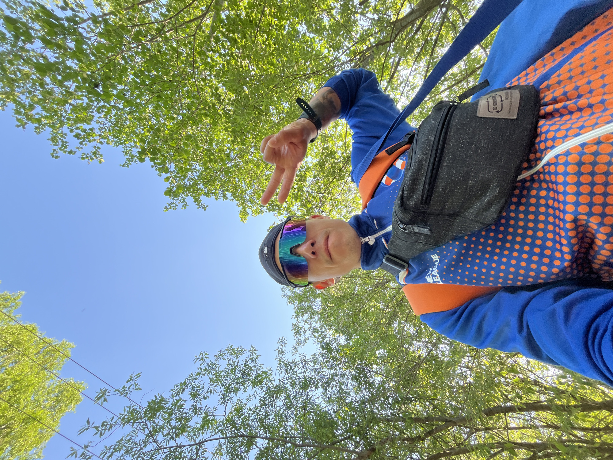 A man is pictured from a low angle, looking down at the camera while making a peace sign. He is wearing a blue and orange patterned sweatshirt, sunglasses with a rainbow sheen, and a black fanny pack. Green foliage and clear blue skies are visible in the background, suggesting an outdoor setting possibly in the city of Chaikovsky, Russia. The scene is bright, suggesting daytime.