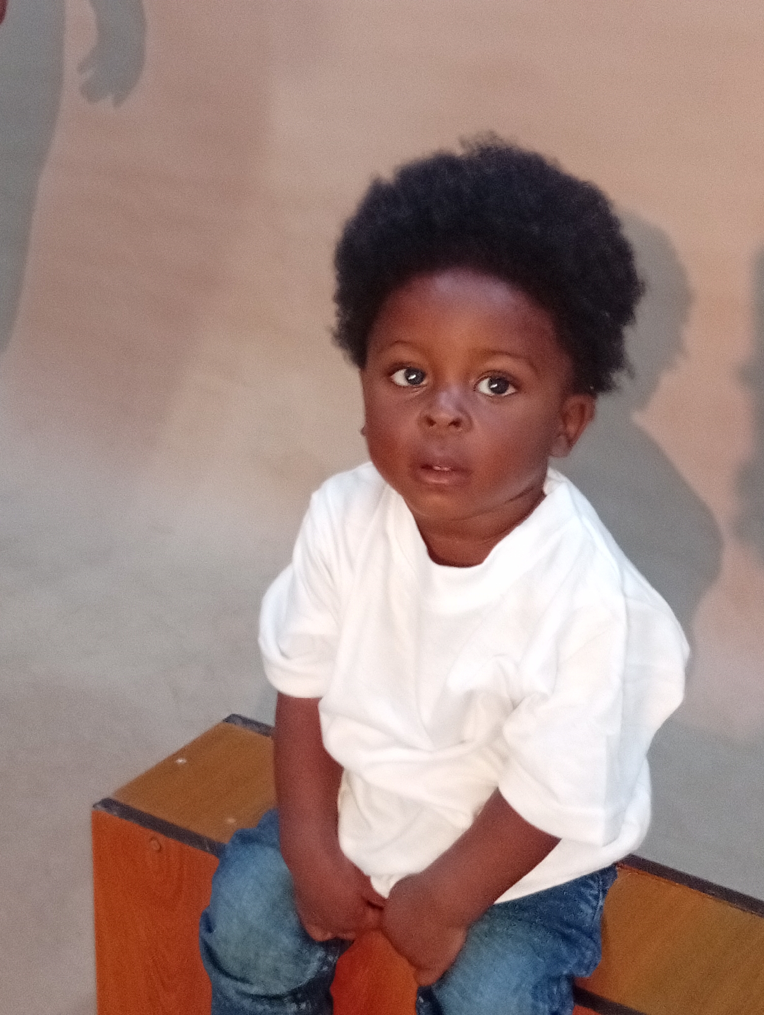 A young Black child with an afro hairstyle is sitting on a wooden surface. The child is wearing a white t-shirt and blue jeans. The child is looking directly at the camera with a neutral expression. The background is a plain, textured wall with a shadow on the left side. The lighting is soft and diffused, suggesting an indoor environment.