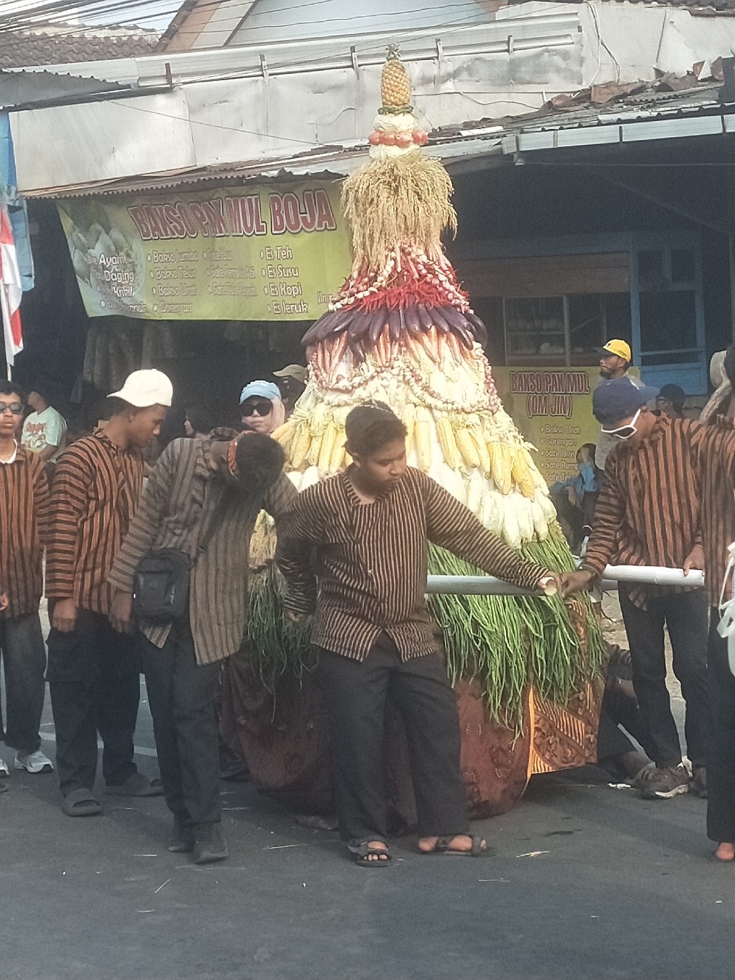 A procession is underway on a paved street in Boja, Indonesia, featuring a large, elaborately decorated float made of vegetables and grains, topped with a pineapple. Several young men, dressed in traditional striped shirts and pants, are carrying the float using poles. The float is adorned with corn, red chilies, and what appear to be eggplants, with long green grass at its base. A banner for "Bakso Pak Mul (Om Jin)" hangs behind the procession. The surrounding environment shows a street scene with buildings and overhead structures, suggesting a public event or festival.