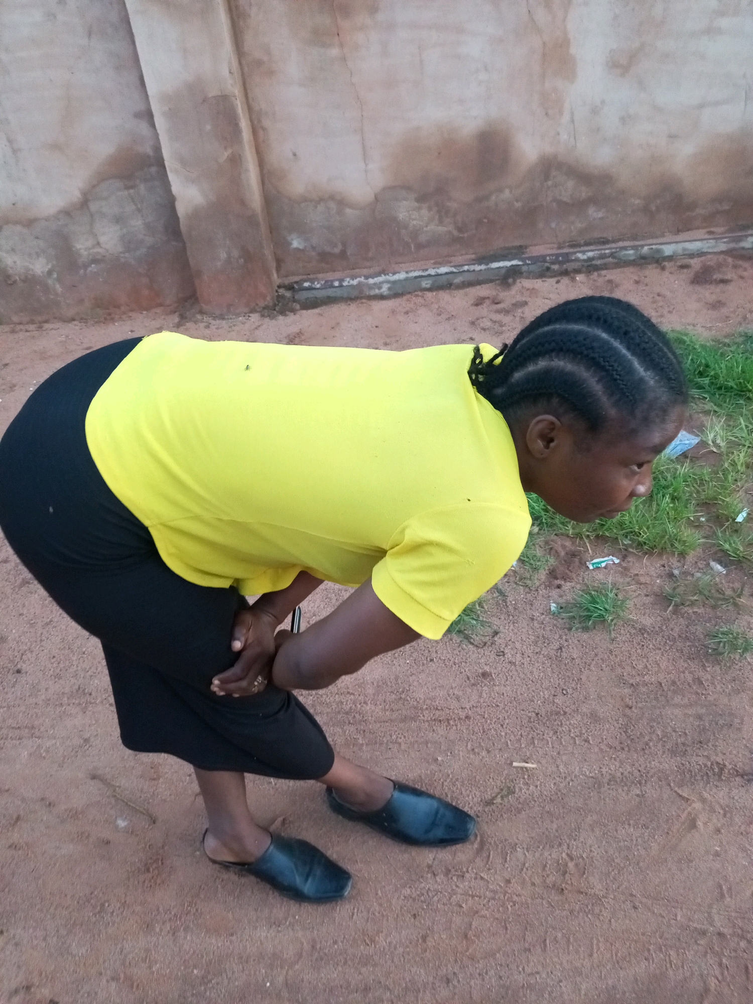 A person wearing a yellow t-shirt and black pants is bent over at the waist, with their head lowered and looking down. They are standing on a dirt or sand ground with some sparse green grass and debris on the right side of the image. A textured, light-colored wall with some discoloration is visible in the background. The person has dark braided hair and is wearing black slip-on shoes. Their hands are clasped in front of their bent knees. The lighting suggests it is daytime.