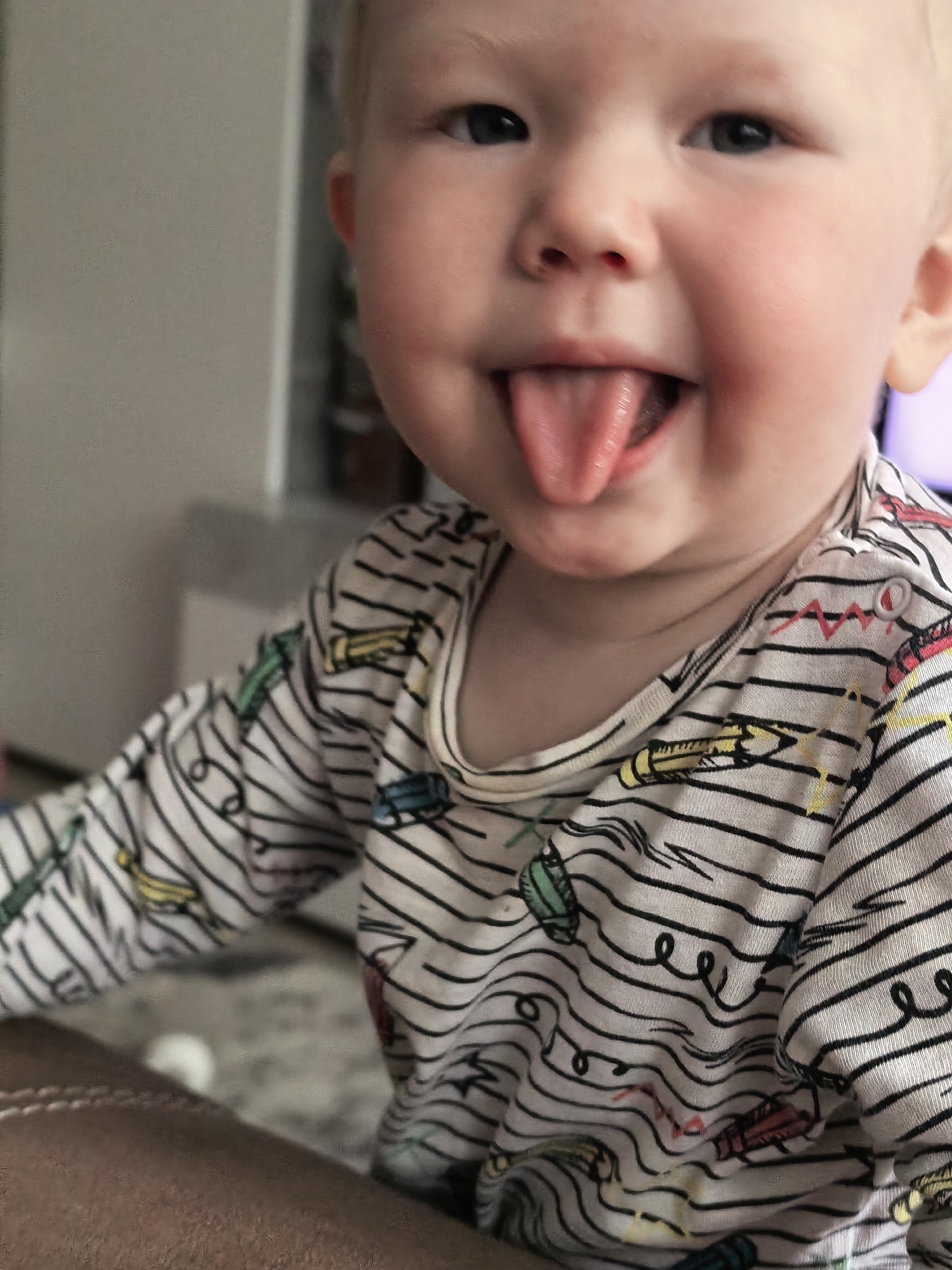 The image, taken in Sterlitamak, Russia, features a close-up, slightly off-center portrait of a baby with fair skin and light-colored hair, primarily visible on the sides of the head. The baby has round, chubby cheeks and dark, expressive eyes that are looking towards the camera. A wide, joyful smile is spread across the baby's face, with the tongue playfully sticking out.
The baby is wearing a long-sleeved shirt with a light background and a pattern of thin, uneven black horizontal stripes. Scattered across the stripes are colorful drawings resembling crayons in shades of yellow, green, red, and blue, along with some squiggly lines and abstract shapes in similar colors. The neckline of the shirt is visible, with a ribbed white trim.
The setting appears to be indoors, likely a home, with a blurred, light-colored background that suggests a wall or furniture. A dark brown, textured object, possibly an armrest of a couch or chair, is visible in the lower left foreground. The lighting is soft and even, highlighting the baby's features. There is no visible text. The overall mood of the image is cheerful and playful, capturing a moment of innocent fun.