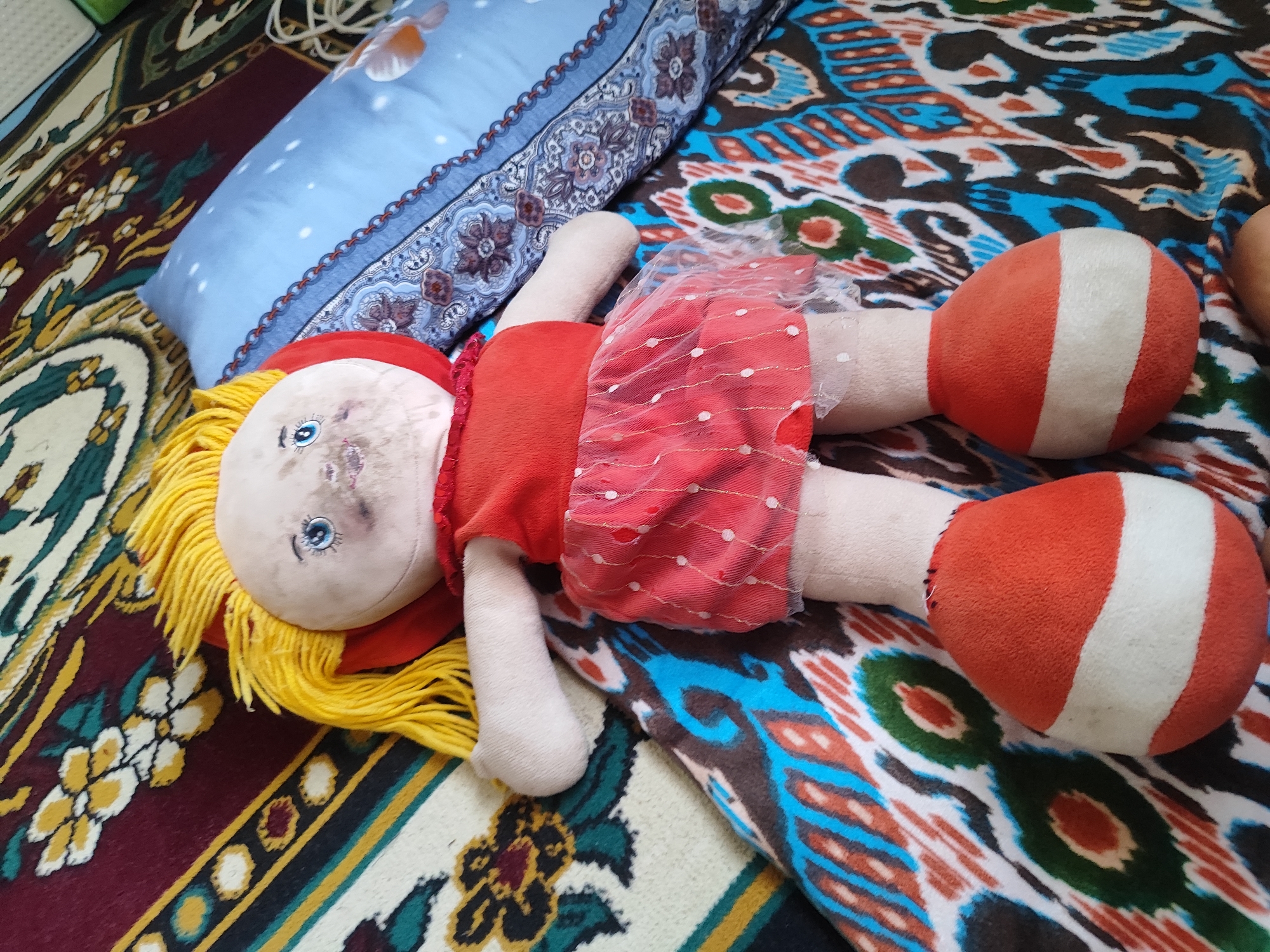 A rag doll with yellow yarn hair and a red dress lies on its back on a patterned blanket. The doll's face has blue eyes and appears smudged. One arm is bent, and its legs are extended. The doll is positioned on a blanket with a colorful, abstract design of blues, browns, greens, and oranges, with a smaller blue pillow adorned with a fish design to its left. The overall scene is likely indoors in the city of Sultonobod, Tajikistan, suggesting a domestic or personal space.
