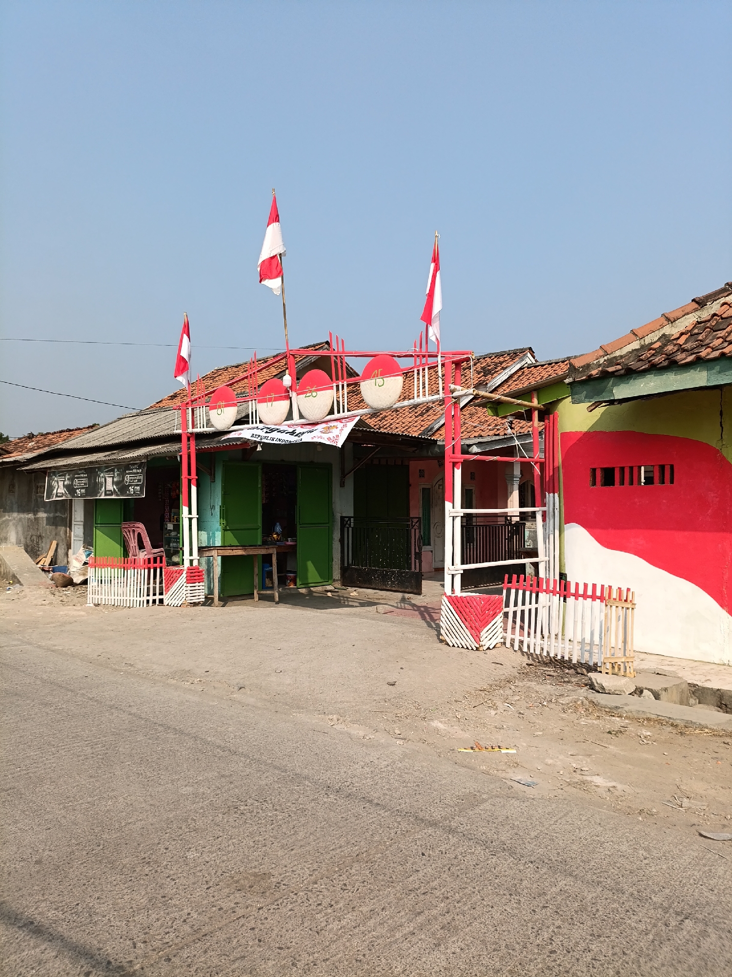 In Cariu, Indonesia, a storefront is decorated with Indonesian flags and festive red and white banners. The building, which appears to be a small shop or market, has green doors and windows. A red and white archway stands in front of the entrance, adorned with circular red and white decorations. A white picket fence, also decorated with red and white elements, surrounds part of the storefront. The building to the right is painted with large red and white wavy stripes. The street in front is paved with concrete, and the sky is clear blue.