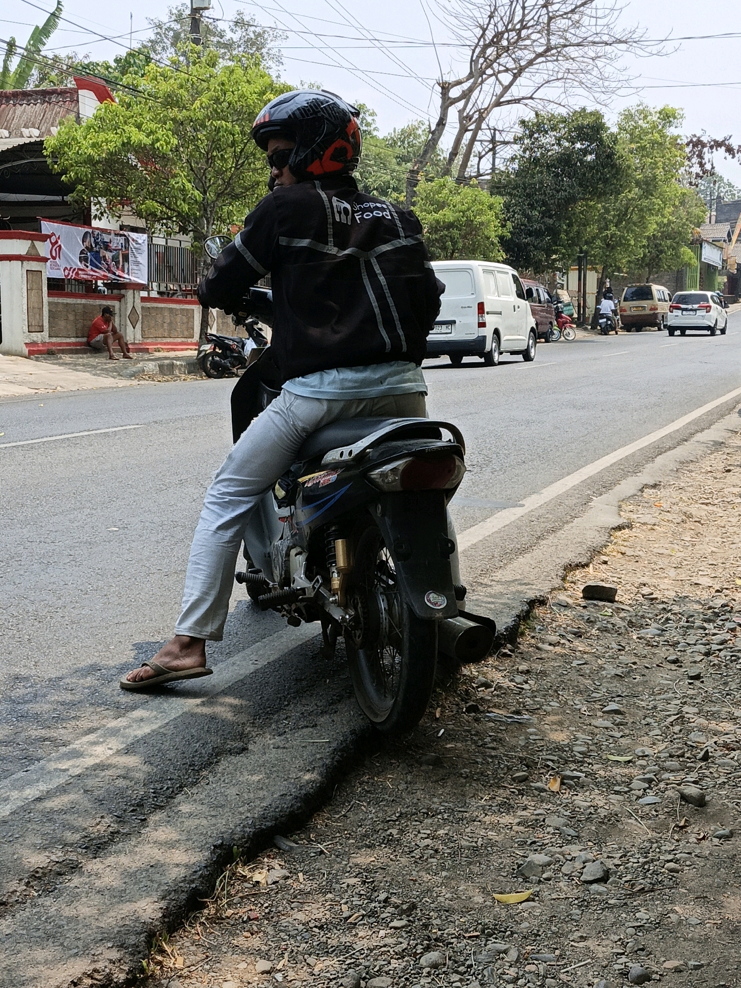 A person wearing a black jacket with reflective stripes and a helmet is sitting on a motorcycle on the side of a paved road in Kuwasen, Indonesia. The motorcycle is black and silver. The person is wearing light-colored pants and flip-flops. A white van and other vehicles are visible on the road in the background. The area appears to be a residential or commercial street with trees and buildings. There is a man sitting on a ledge on the left side of the image. The road has some damage and gravel on the side.