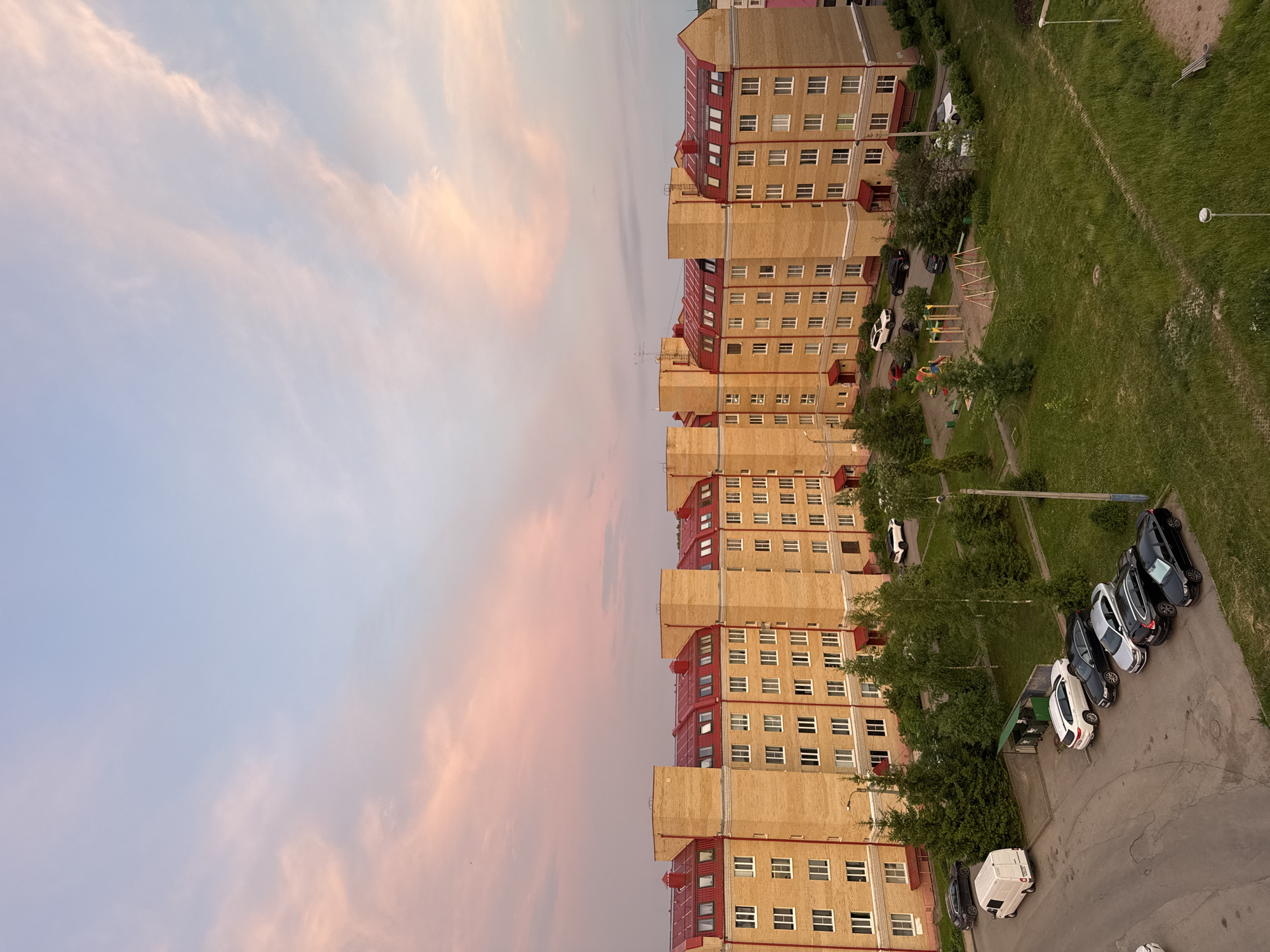 An aerial perspective captures a residential apartment complex in Kirishi, Russia, bathed in the soft, pastel hues of a sunset. The building, constructed from pale yellow brick with distinctive red-tiled roofs and visible dormer windows, stretches horizontally across the frame. Numerous white-framed windows punctuate the facade, suggesting multiple stories and apartments.
In front of the building, a paved area serves as a parking lot, containing several vehicles. These include a white van, a white SUV, a black SUV, a silver sedan, and two black sedans, all parked in an orderly fashion. Adjacent to the parking area and the building, a patch of green lawn is interspersed with several evergreen trees and a small children's playground featuring swings and colorful play structures.
Further away, more trees and green spaces are visible, contributing to the suburban feel of the scene. The sky above is a canvas of pale blue and soft pink clouds, indicative of twilight. The overall atmosphere is tranquil and domestic, depicting a typical evening in a Russian residential area.
