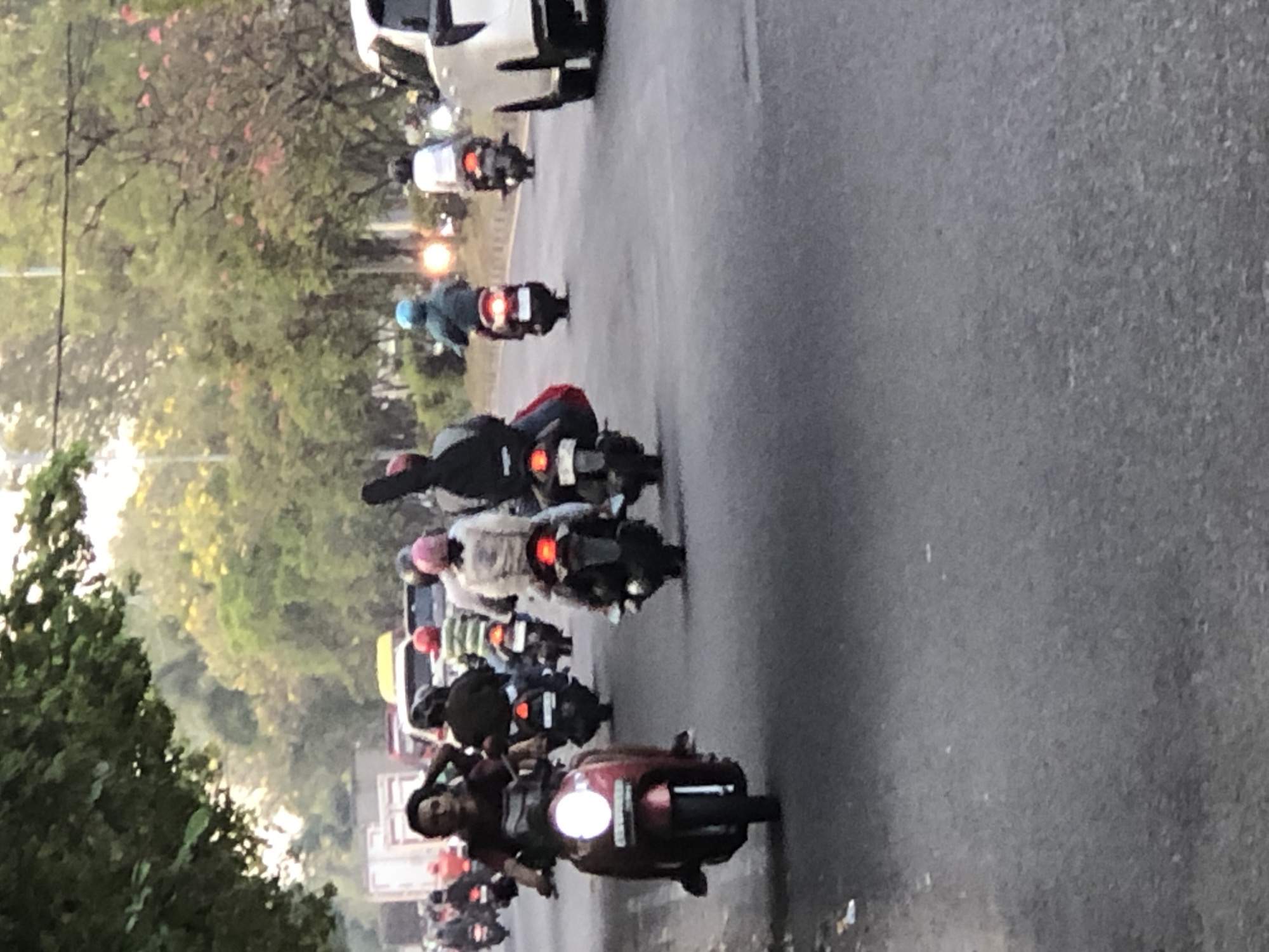 The image depicts a street scene in Sumorame, Indonesia, characterized by heavy motorcycle traffic. The road is asphalt and appears to be an urban thoroughfare with trees lining the sides. In the foreground, a person on a red scooter with its headlight illuminated is prominently featured. Several other individuals on motorcycles are visible further down the road, some carrying passengers. The overall impression is one of daily commute or travel within the city. The lighting suggests it may be late afternoon or early evening.