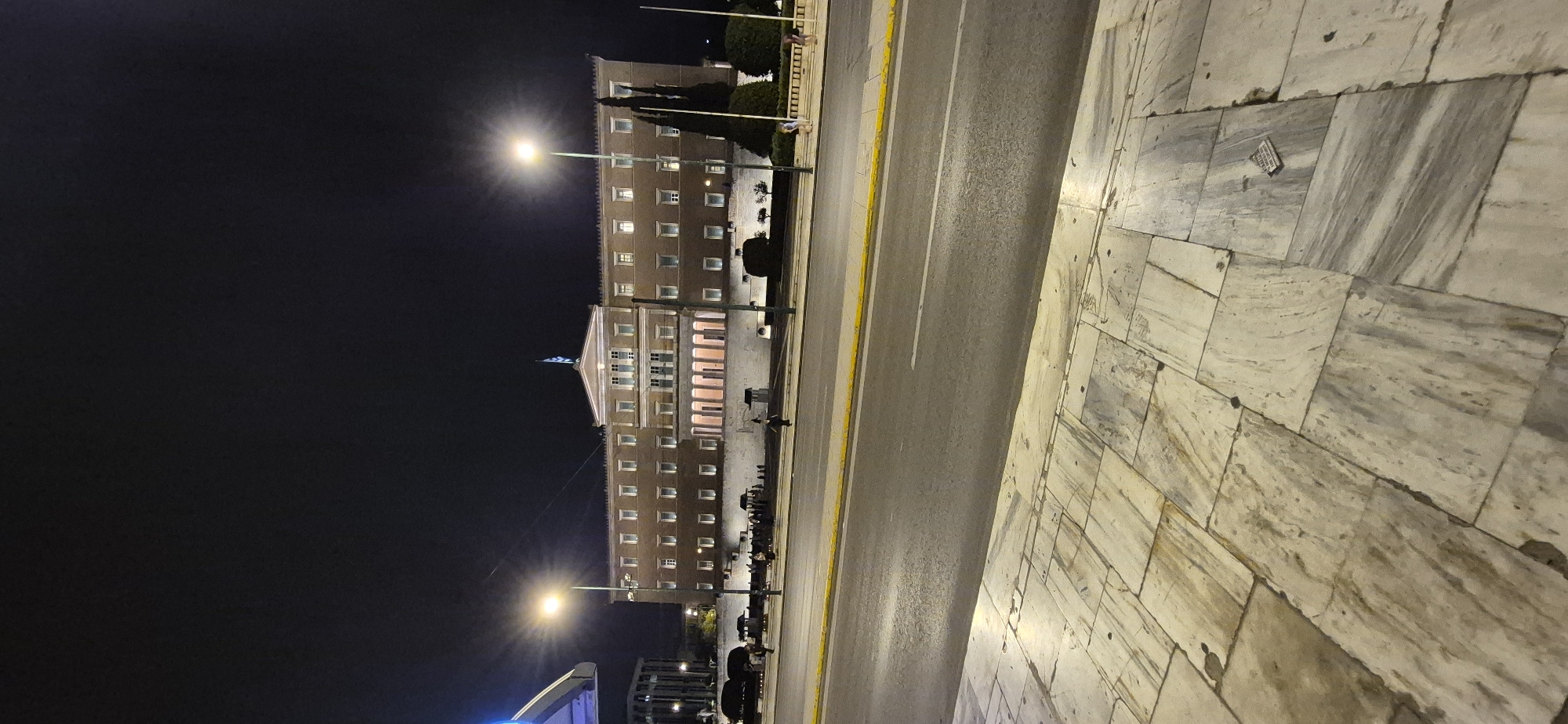 This media file depicts a nighttime scene in Athens, Greece, likely near a significant building. The image is oriented vertically, with the ground on the right and the sky on the left. A large, multi-story building with many illuminated windows dominates the left side of the frame. The architecture appears classical, with a pedimented entrance and columns visible. A Greek flag is mounted on the building.
In front of the building, there is a street with at least two lanes of traffic, marked by a yellow line. Streetlights are present, casting a warm glow on the street and sidewalk. The sidewalk to the right is paved with large, marble-like tiles, showing some wear and tear.
There are a few people visible in the scene, mostly silhouettes against the lights of the building and the street. They appear to be standing or walking, suggesting a casual evening atmosphere. Some trees and bushes are also visible, adding to the urban landscape.
The time of day is clearly night, indicated by the dark sky and the artificial lighting. The weather appears clear. The marble paving and the architectural style of the building are strong cues for its location in Athens. No discernible text is visible in the image. The overall mood is calm and urban, with the lights creating a somewhat serene ambiance.
