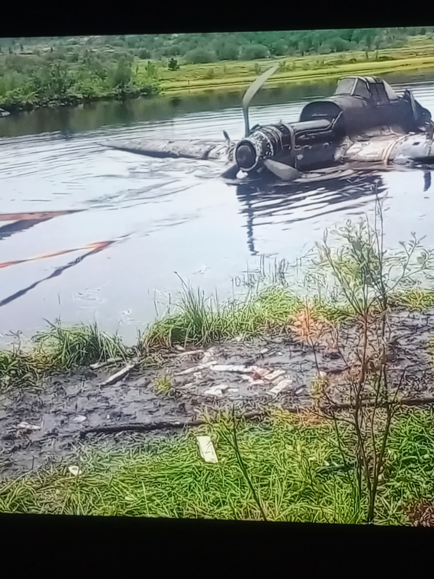 The image shows the wreckage of an airplane partially submerged in water, likely a river or lake. The aircraft, a dark-colored propeller plane, appears to be an older model. Its fuselage is mostly underwater, with the tail section and one wing visible above the surface. The propeller is intact and partially submerged. The scene is set against a backdrop of lush green trees and vegetation, suggesting a natural, possibly rural, environment. The water is rippled, and the sky is not visible, making it difficult to determine the time of day or weather conditions. The foreground shows a muddy bank covered in grass and debris, with some dry branches and what appears to be trash. There is a hint of orange material, possibly a rope or strap, extending from the aircraft into the water on the left side of the image. The overall impression is one of a recent crash or abandonment in a remote natural setting. No people are visible in the image. There is no discernible text.