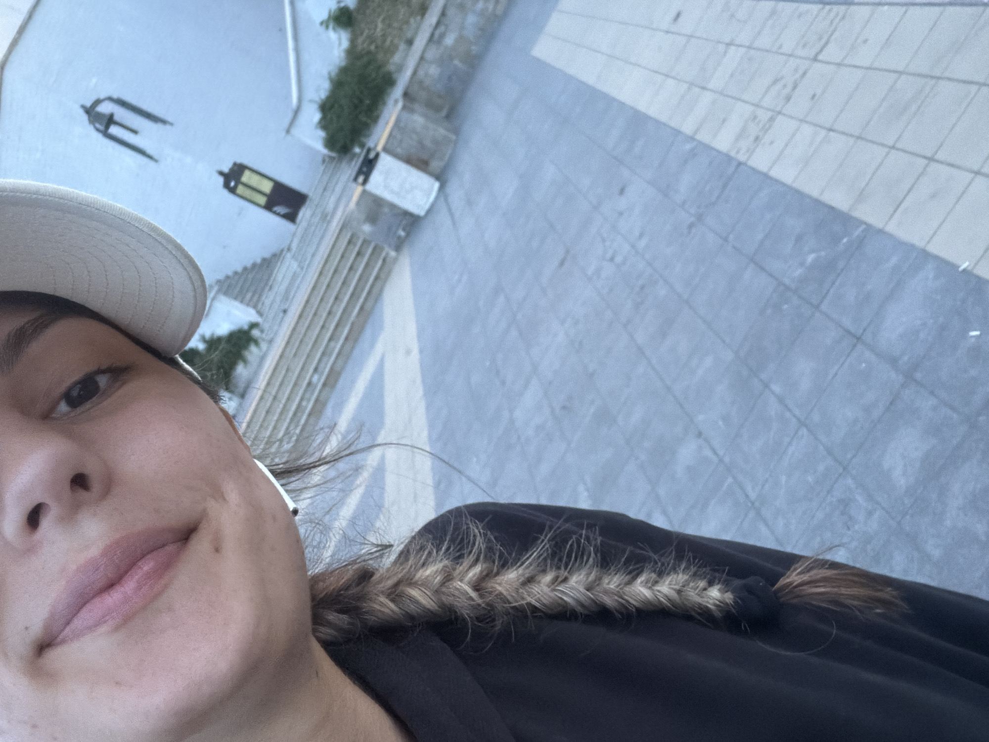 The image shows a close-up selfie of a young person with fair skin and dark hair, wearing a black hoodie and a light-colored baseball cap. Their hair is styled in a braid that hangs over their left shoulder. The background is out of focus but appears to be an outdoor setting with tiled flooring and a white building with a bell and a dark-colored door. The overall lighting suggests it is daytime. The image does not contain any specific elements that would allow for a definitive location within Burgas, Bulgaria.